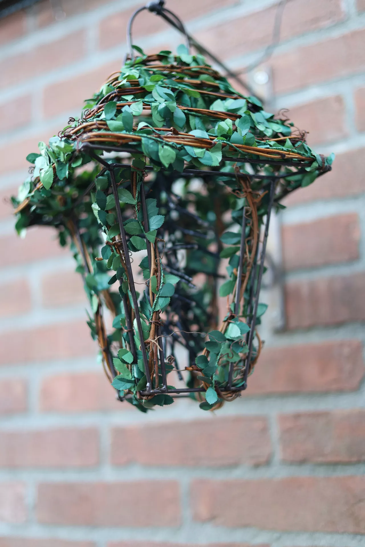 A unique outdoor display showcases garden-themed lantern summer decor ideas hanging against a classic red brick wall. The creative lantern with ivy features an open, dark metal wireframe cage shaped like a traditional street lantern. Thick wooden vines and lush green ivy leaves are intricately wrapped around the metal bars, completely covering the top cap and trailing down the sides of the structure. The plant-covered lantern hangs from a metal mount, bringing a natural, botanical touch to an outdoor brick patio or garden wall.