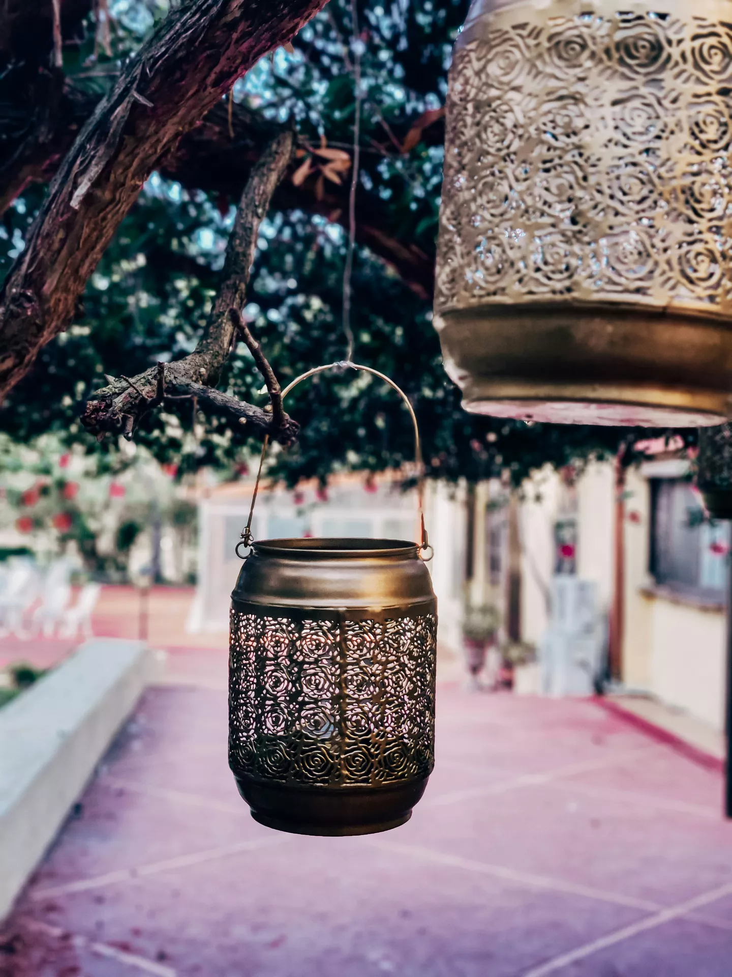 An atmospheric outdoor setting highlights beautiful lantern summer decor ideas suspended from the branches of a large tree. The display features two ornate metallic lanterns crafted from brass or gold-toned metal with intricate, lace-like filigree cutouts across their cylindrical bodies. One lantern hangs prominently in the lower-middle foreground from a thin metal wire handle tied to a branch, while the second larger lantern hangs closer to the top right corner of the frame. In the softly blurred background, a warm backyard courtyard with a reddish-pink tiled patio and residential buildings creates a relaxed summer evening vibe.