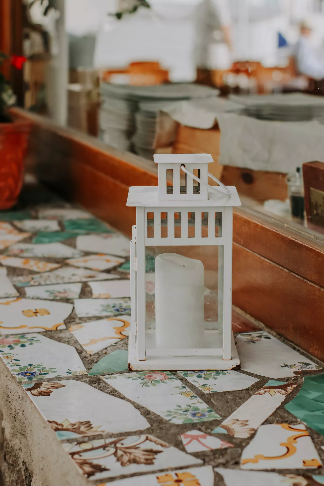 A charming outdoor setting highlights rustic lantern summer decor ideas arranged on a unique mosaic stone surface. A clean white lantern with pillar candle sits as the centerpiece, featuring a simple rectangular frame with glass panels and a small tiered top with ventilation slots. The lantern rests on a ledge made of mismatched, colorful broken ceramic tiles adorned with floral patterns. In the softly blurred background, a warm wooden window sill and reflections of a cozy cafe or restaurant interior enhance the bright, Mediterranean holiday atmosphere.