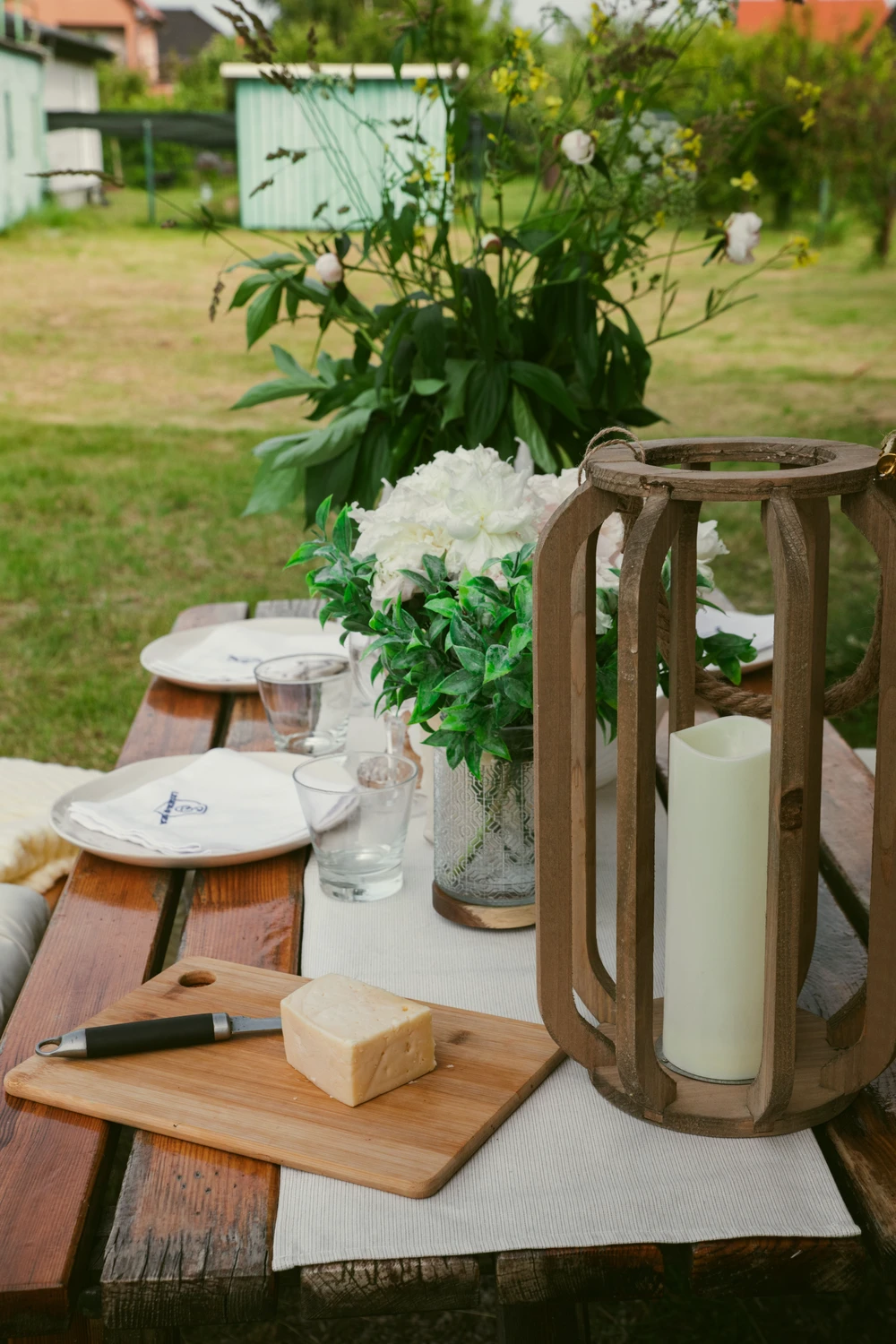 This description for lantern-summer-decor-ideas-wooden-lantern.webp captures an inviting outdoor dining setup featuring rustic lantern summer decor ideas on a wooden garden table. In the foreground, a large minimalist wooden lantern crafted from curved vertical slats holds a tall white pillar candle on top of a light-colored table runner. Right next to it, a small wooden cutting board displays a block of cheese and a small knife. The festive table arrangement continues with clear drinking glasses, white plates with napkins, and a glass vase filled with beautiful white peonies and green foliage. In the blurred background, a lush green lawn and a small mint-green garden house complete the peaceful backyard atmosphere.