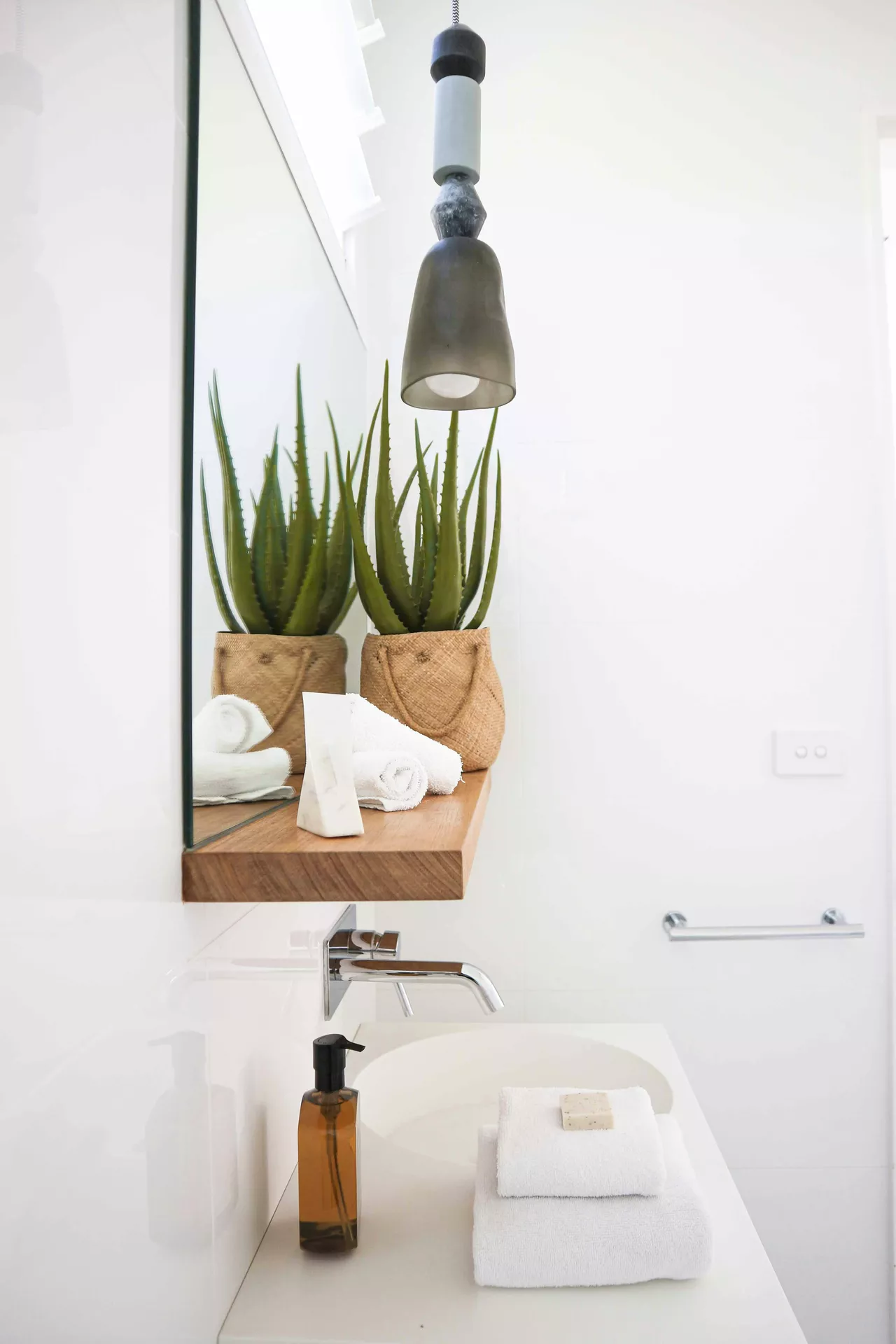 This description for neutral-bathroom-decor-ideas-aloe-vera-plant.webp captures a bright and modern vanity area showcasing clean neutral bathroom decor ideas. A vibrant aloe vera plant sits in a woven jute basket on a floating wooden shelf alongside a small marble accessory and rolled white hand towels, all reflected beautifully in a large frameless mirror. Below the shelf, a contemporary white basin vanity features a sleek chrome wall-mounted faucet, an amber glass soap dispenser, and a stack of folded white towels holding a bar of natural soap. A translucent dark grey pendant light hangs gracefully from above, completing the serene and minimalist bathroom aesthetic against the crisp white walls.