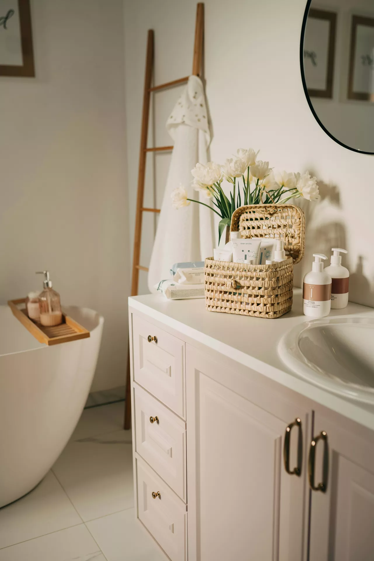 This description for neutral-bathroom-decor-ideas-basket.webp highlights a beautifully organized vanity area displaying elegant neutral bathroom decor ideas. On the smooth white countertop of a light beige vanity cabinet with gold handles, an open woven straw basket with a lid functions as a stylish storage container filled with various skincare tubes. Next to the container stand two minimalist white and brown lotion dispenser bottles, while a vase filled with delicate white tulips adds a fresh touch behind them. In the soft-focus background, a freestanding white bathtub with a wooden caddy tray, a wooden decorative ladder holding a white hooded towel, and a round black-framed mirror on the wall complete the peaceful, spa-like atmosphere.