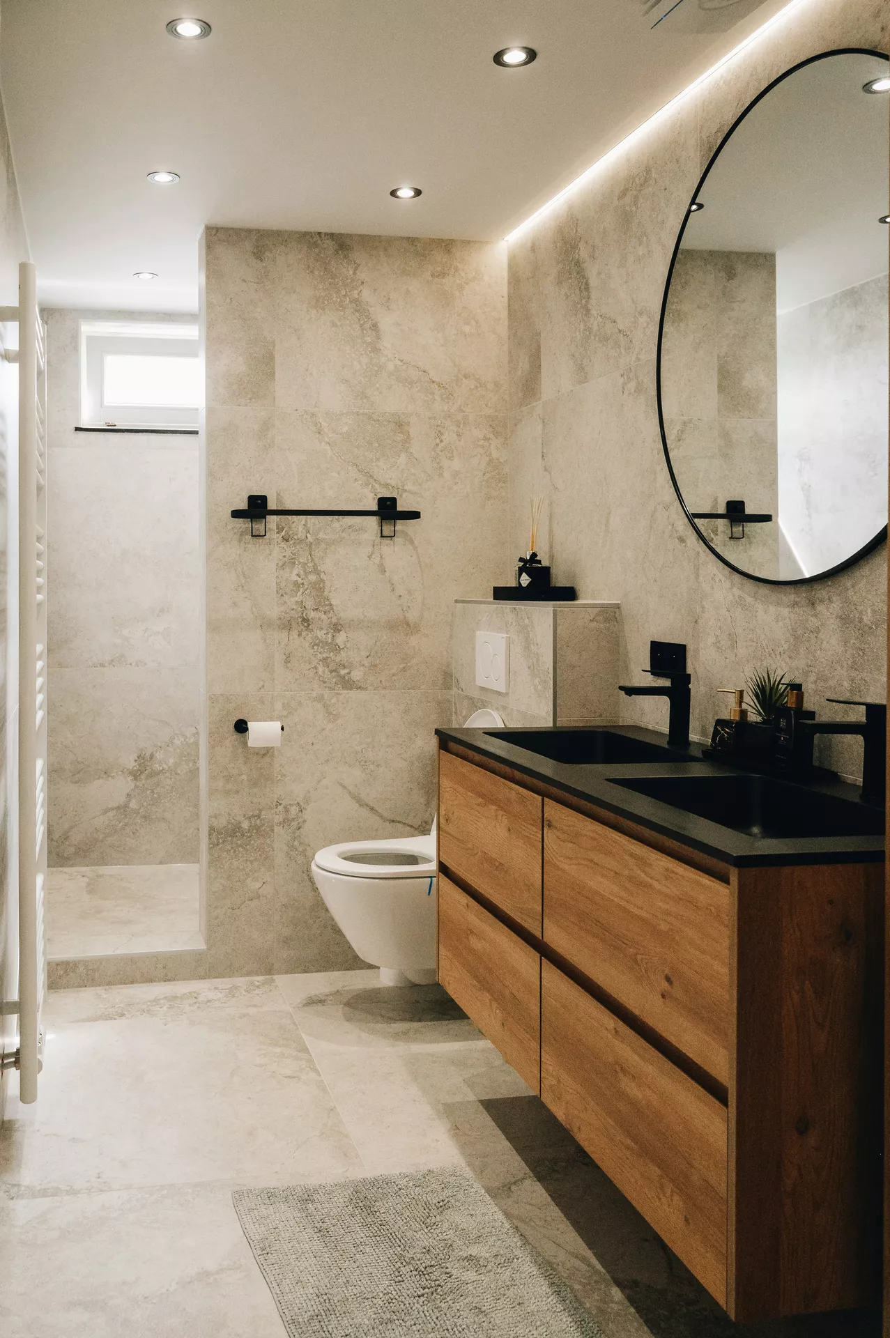 This description for neutral-bathroom-decor-ideas-black-fixtures.jpg showcases a luxurious and contemporary bathroom design featuring sophisticated neutral bathroom decor ideas. The space is wrapped in large beige marble-look porcelain tiles on the walls and floor, creating a cohesive, spa-like feel. A floating wooden double vanity with a dark charcoal countertop and dual undermount sinks takes center stage, accented by sleek matte black fixtures including two modern black faucets and a matching black towel bar. A large oval mirror with a thin black frame hangs above the vanity, illuminated by hidden LED strip lighting along the ceiling edge and recessed spotlights. In the background, a wall-hung white toilet sits near a seamless walk-in shower area equipped with a white towel radiator on the left wall, perfecting this elegant and minimalist interior.