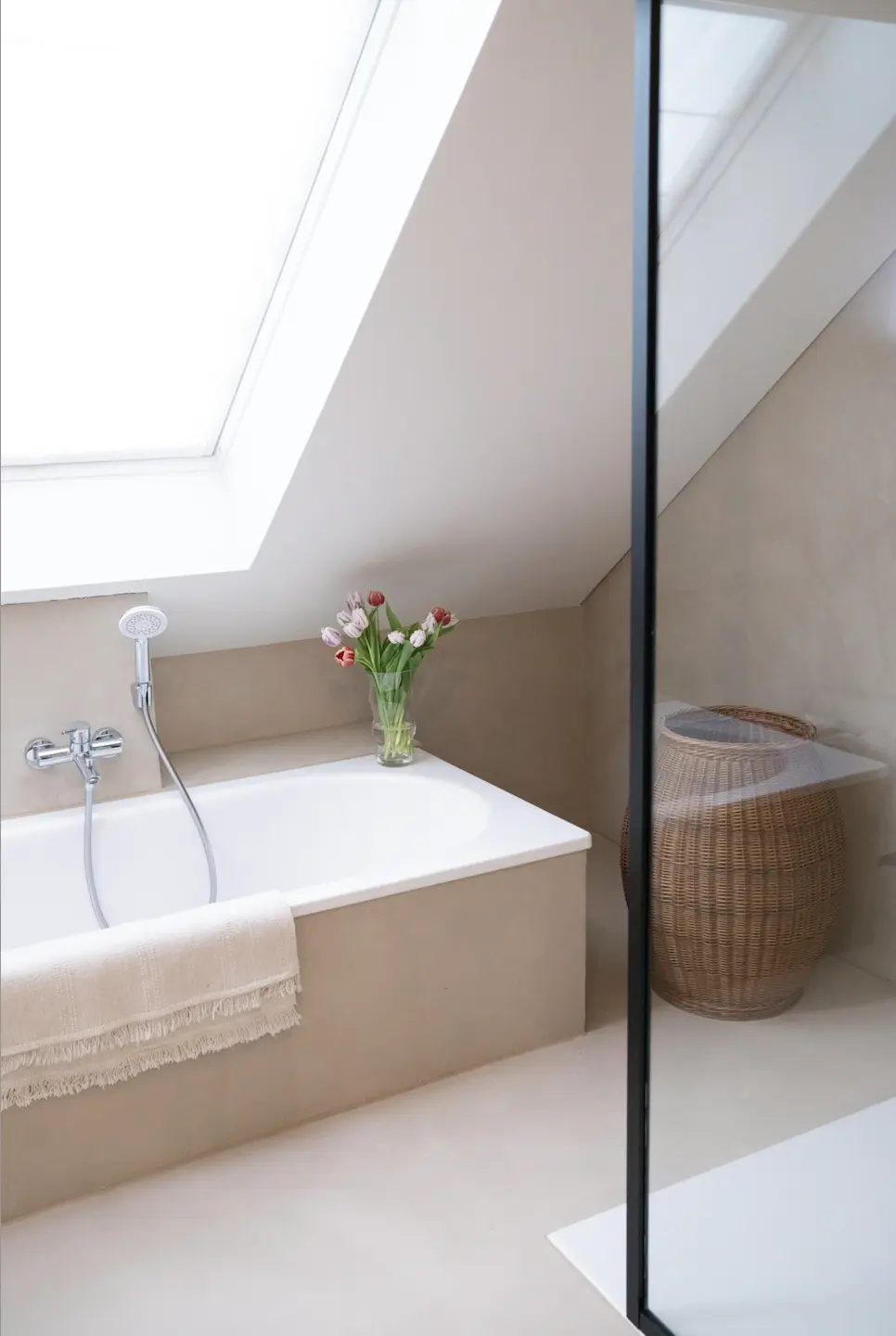 This description for neutral-bathroom-decor-ideas-fresh-flowers.webp captures a bright attic bathroom showcasing minimalist neutral bathroom decor ideas. A modern built-in white bathtub with warm beige tiling features a sleek chrome faucet and handheld showerhead, with a fringed beige towel draped neatly over the side. Positioned on the corner of the tub is a clear glass vase filled with lovely pink and red fresh flowers, adding a natural pop of color under a large slanted skylight window that floods the space with natural light. To the right, a large woven wicker laundry basket stands in the corner, partially seen through a black-framed glass shower screen, completing the serene and organic aesthetic.
