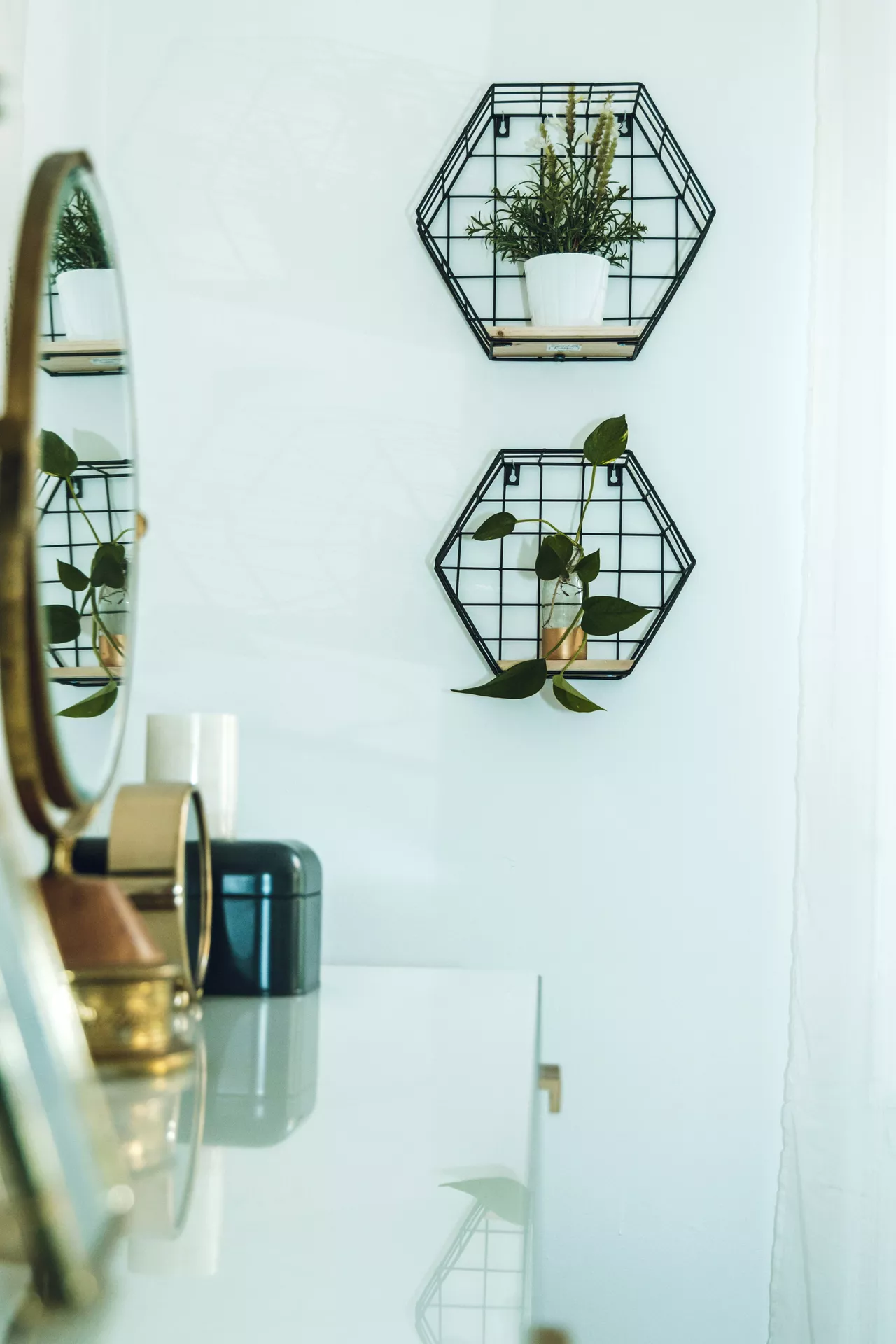 This description for neutral-bathroom-decor-ideas-geometric-storage-unit.webp highlights a bright, crisp vanity wall featuring modern neutral bathroom decor ideas. Mounted on the clean white wall is a stylish geometric storage unit consisting of two black wireframe hexagon shelves with light wood bases. The top shelf holds a small white pot with delicate greenery, while the lower shelf displays a single leafy green plant cutting inside a small glass vase. In the foreground, a sleek white vanity tabletop reflects the arrangement, showcasing blurred gold accents, small storage containers, and a vanity mirror on the left side that captures a reflection of the botanical wall decor.