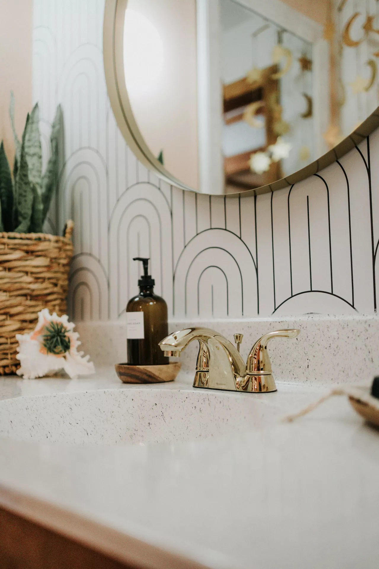 This description for neutral-bathroom-decor-ideas-gold-colored-fixtures.webp highlights a stylish and modern bathroom vanity area showcasing creative neutral bathroom decor ideas. A polished, elegant gold-colored faucet sits centered on a light-colored speckled countertop next to a white undermount sink. An amber glass soap bottle rests in a small wooden dish nearby. The background features a striking black and white geometric wallpaper pattern of repeating arches, partly covered by a large round mirror with a thin gold frame. On the left side of the vanity, a snake plant in a woven wicker basket and a large decorative seashell with a small succulent inside add natural texture to the serene arrangement.