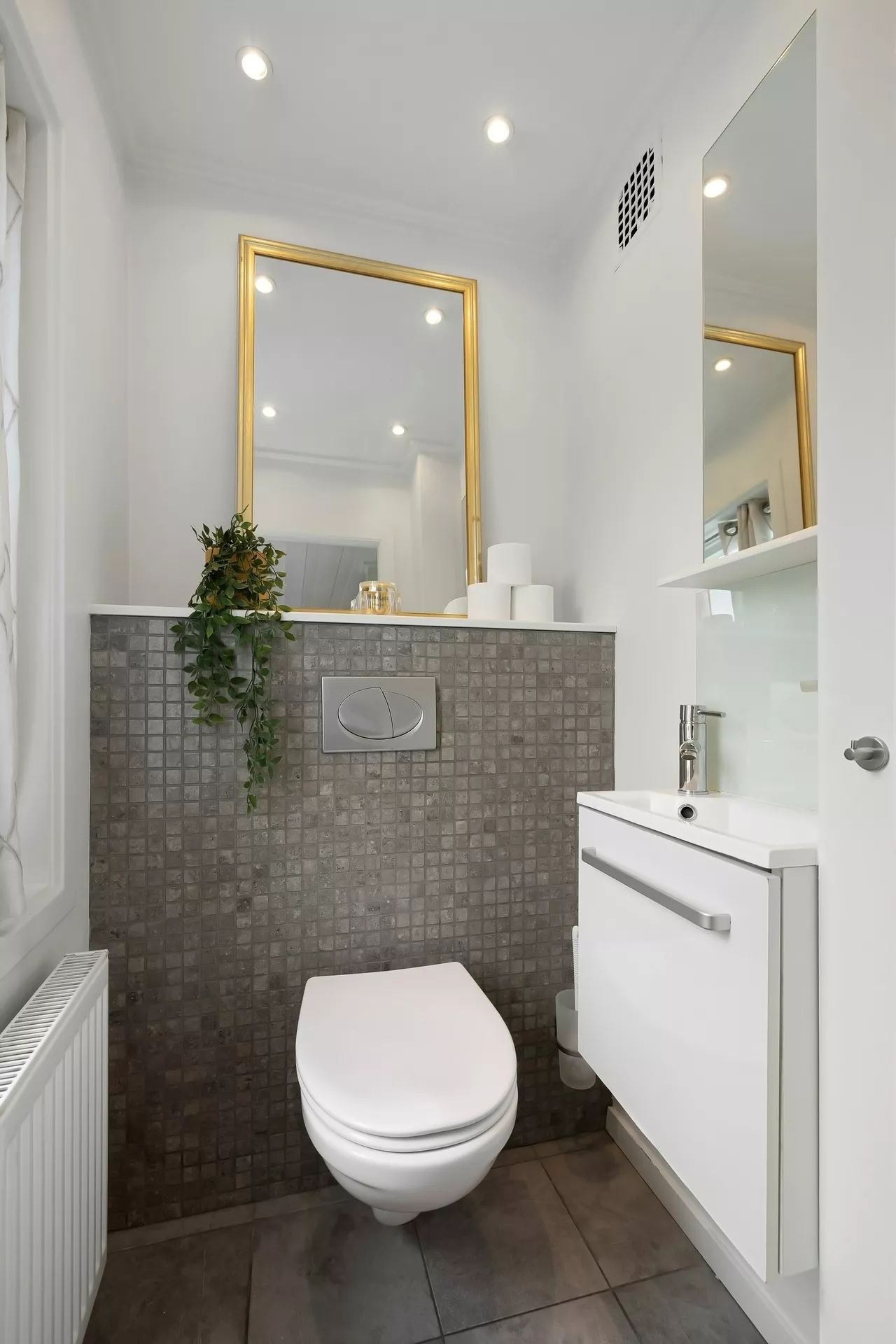This description for neutral-bathroom-decor-ideas-golden-mirror.webp illustrates a compact and modern powder room filled with clean neutral bathroom decor ideas. The main feature is a large golden mirror with a metallic frame resting on a shelf above a wall-hung toilet. The accent wall behind the toilet is covered in small, square mosaic tiles in shades of grey, while the surrounding walls are painted a crisp white. On the right side, a compact white floating vanity sink with a silver chrome faucet sits under another vertical frameless mirror. A trailing green plant in a pot sits on the ledge next to toilet paper rolls, adding a touch of natural color under bright ceiling spotlights.