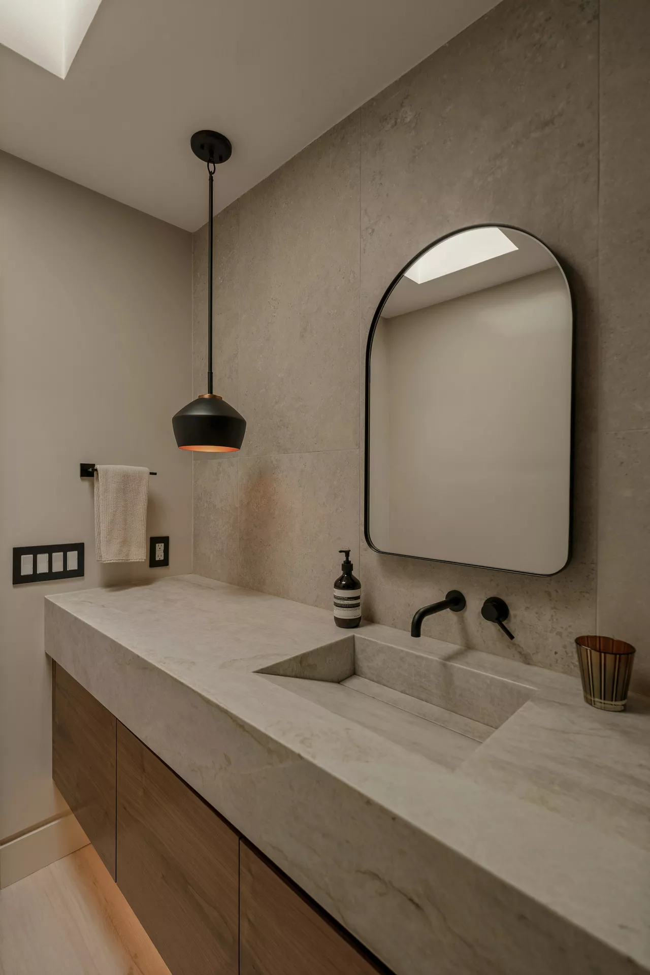 This description for neutral-bathroom-decor-ideas-hanging-lamp.webp highlights a sophisticated, minimalist vanity space featuring contemporary neutral bathroom decor ideas. A sleek black hanging lamp with a warm metallic interior glows gently on the left, suspended over a large integrated stone sink and countertop. The textured light-grey tiled wall behind the vanity is accented by a wall-mounted matte black faucet and a matching black-framed arched mirror. Underneath the stone countertop, floating wooden cabinetry provides a warm contrast, complete with a clean white side wall holding a black towel rack and matching electrical switches.
