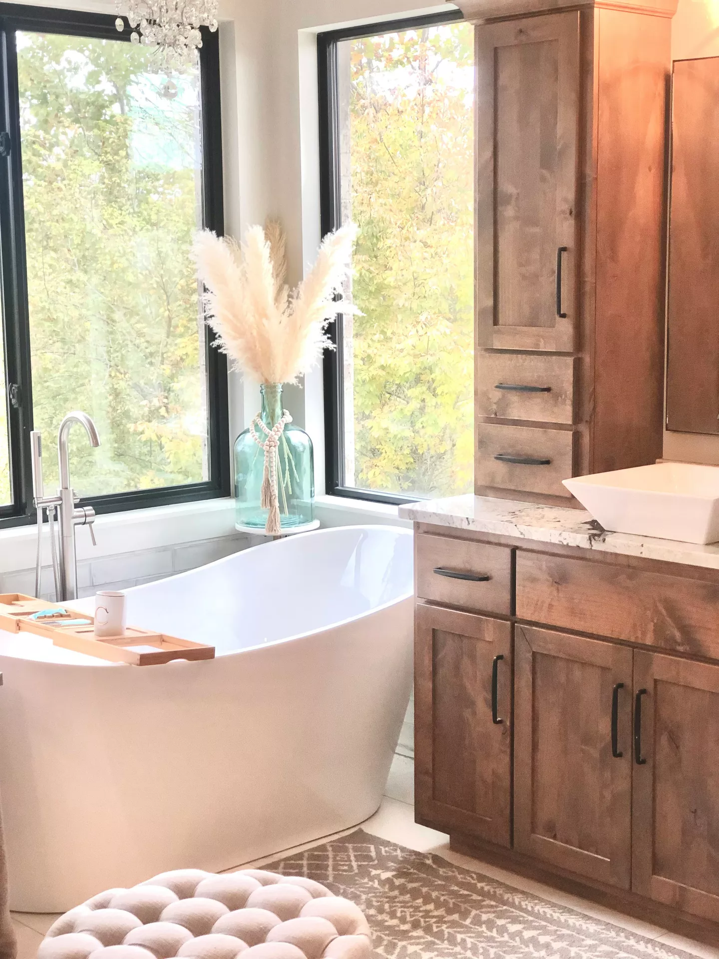 This description for neutral-bathroom-decor-ideas-pampas-grass.jpg showcases a spacious and elegant washroom filled with warm neutral bathroom decor ideas. In the center, a large freestanding white oval bathtub sits next to large black-framed windows that reveal a soft background of autumn trees. Fluffy stalks of beige pampas grass are displayed beautifully inside a large translucent turquoise glass jug on a small pedestal behind the tub, decorated with a string of wooden beads. To the right, a rich brown wooden vanity cabinet with black handles features a white vessel sink on a light marble countertop. A wooden bath caddy tray rests on the bathtub, while a tufted beige ottoman and a patterned area rug on the floor complete the cozy, spa-like atmosphere.