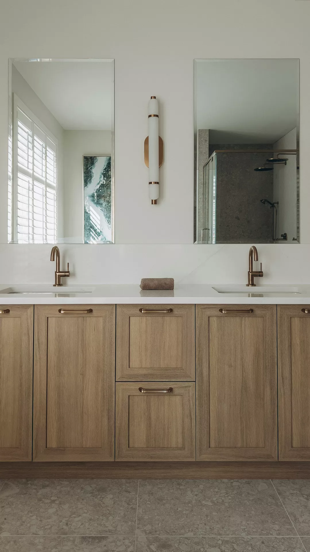This description for neutral-bathroom-decor-ideas-pretty-bathroom-cabinets.webp highlights a symmetric and elegant double vanity space featuring modern neutral bathroom decor ideas. A large light wood vanity unit is fitted with pretty bathroom cabinets and drawers with thin metallic handles. On top, a solid white countertop holds two rectangular undermount sinks, each paired with a contemporary brushed bronze or gold-toned faucet. Two tall, frameless rectangular mirrors hang on the white wall above each sink, reflecting a window with shutters on the left and a stone-tiled walk-in shower on the right. A sleek, vertical cylindrical wall sconce light with metallic accents is mounted centered between the two mirrors, completing the calm, upscale bathroom aesthetic above the grey stone-textured floor tiles.