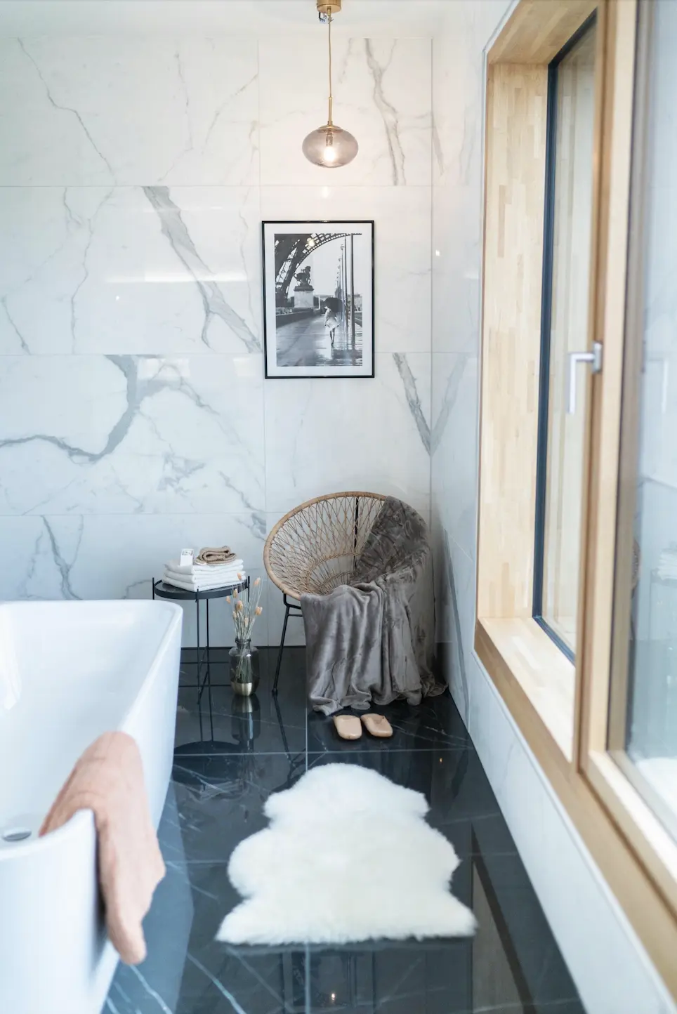 This description for neutral-bathroom-decor-ideas-seating-area.webp showcases a luxurious, spa-like washroom corner designed with contemporary neutral bathroom decor ideas. A polished black marble floor creates a bold contrast against large, glossy white marble wall tiles. In the corner, a cozy seating area is established with a round woven wicker chair draped with a plush grey blanket, paired with a small pair of slippers on the floor and a white faux-fur sheepskin rug in the foreground. Above the chair, a black-and-white framed art piece hangs under a delicate glass globe pendant light. On the left, a small black side table holds a neat stack of white towels, positioned next to a large freestanding white bathtub with a soft peachy-pink towel draped over the rim, while a large wood-framed window on the right fills the space with natural light.