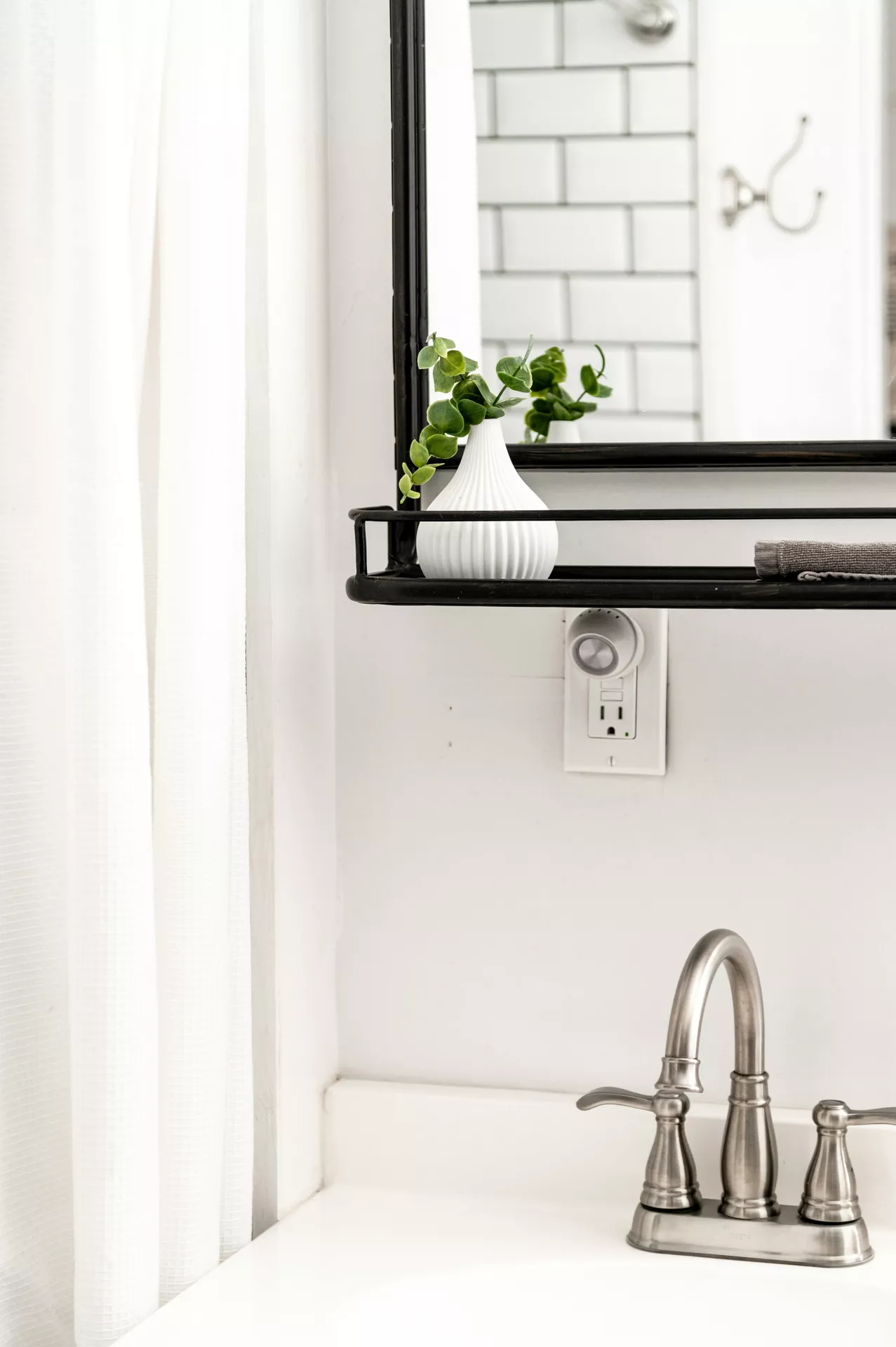 This description for neutral-bathroom-decor-ideas-small-white-vase.webp highlights a clean and bright vanity area showing minimalist neutral bathroom decor ideas. In the lower right corner, a brushed nickel faucet sits on a white countertop below a crisp white wall. Attached to the wall is a black metal wire shelf integrated with the base of a black-framed mirror. On the shelf rests a small white vase with textured vertical ridges, holding a few fresh green eucalyptus sprigs that are reflected in the glass. The mirror also captures a glimpse of a classic white subway-tiled wall in the background, while a soft white fabric shower curtain drapes on the left side, completing the bright and clean aesthetic.