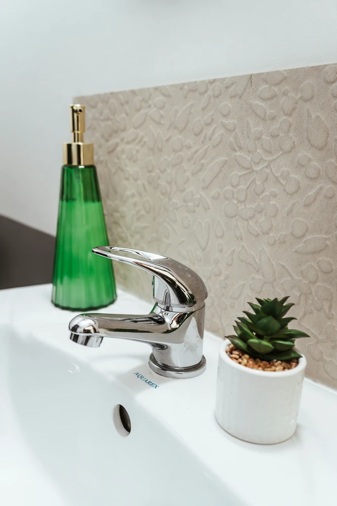 This description for neutral-bathroom-decor-ideas-succulents.webp captures a close-up of a modern vanity station showcasing fresh neutral bathroom decor ideas. A polished silver chrome faucet is mounted onto a clean white ceramic basin. To the right of the tap, a small white ceramic pot holds green succulents nestled in tiny brown pebbles. In the background, a beautiful beige backsplash tile features an embossed, textured floral pattern, adding subtle depth to the washroom setting. On the left side of the sink, a tall green glass soap dispenser with a gold pump adds a delicate pop of color to the otherwise calm and organic vanity space.