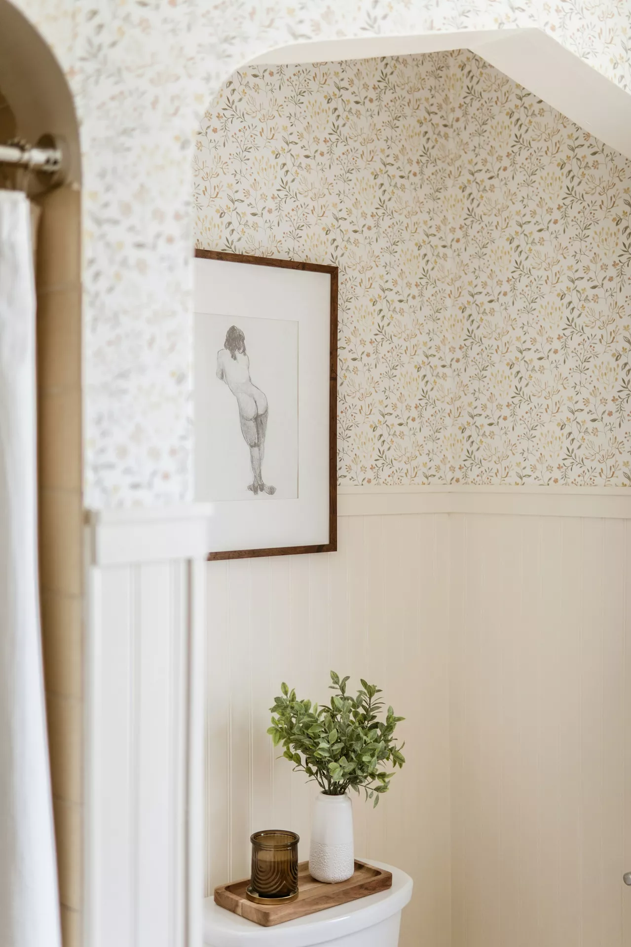 This description for neutral-bathroom-decor-ideas-tray-with-decor.webp highlights a cozy and charming corner filled with vintage-inspired neutral bathroom decor ideas. Above the toilet tank, the upper wall is covered in a delicate floral wallpaper pattern featuring muted yellow and green leaves, separated from the cream-colored vertical shiplap wainscoting below by a thin wood trim. A framed black and white sketch of a female figure hangs on the wall, adding an artistic touch. Resting on the white toilet tank is a small rectangular tray with decor, crafted from natural wood and holding a textured white vase filled with fresh green leafy stems alongside a small, ribbed amber glass cup that beautifully catches the soft light.