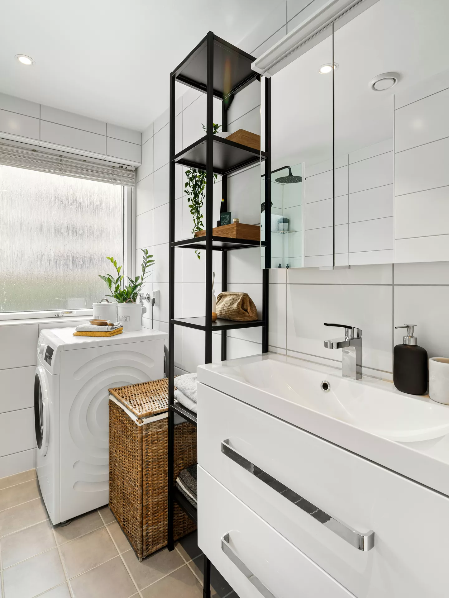 This description for neutral-bathroom-decor-ideas-vertical-shelf.webp illustrates a bright, organized bathroom filled with functional neutral bathroom decor ideas. In the foreground, a modern white vanity unit with large chrome handle drawers holds a white sink with a sleek silver faucet and a dark brown soap dispenser. Mounted above the sink is a mirrored medicine cabinet that reflects the tiled space. Centered in the room stands a tall black metal vertical shelf used for stylish organization, holding folded towels, wooden storage boxes, a woven pouch, and a trailing green houseplant. In the background under a large window, a white washing machine is positioned next to a tall wicker laundry basket, creating a clean, seamless look against the large white wall tiles and light beige floor tiles.