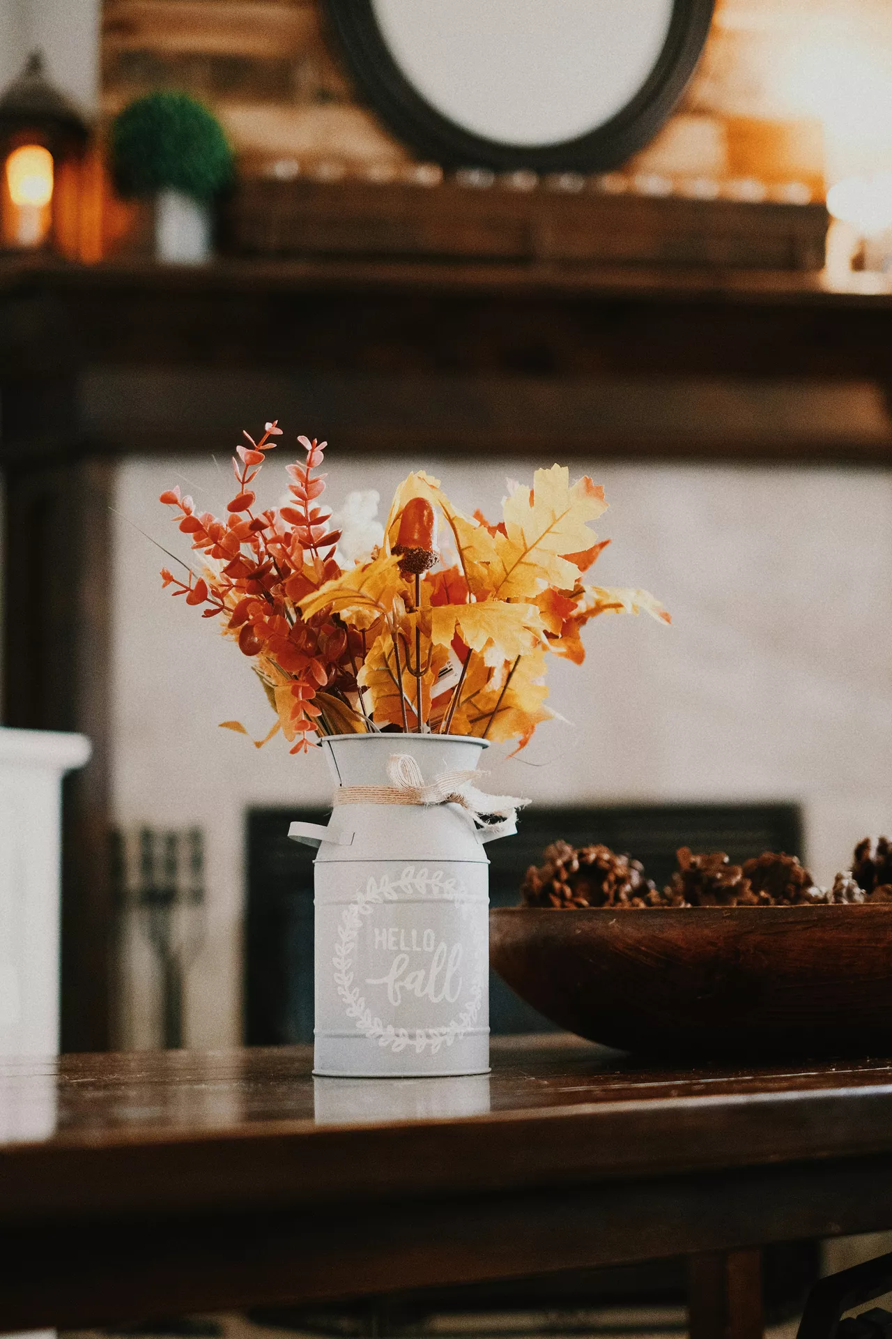 This image showcases cozy rustic fall table decor ideas on a polished dark wooden surface. In the center stands a light blue-grey metal milk jug vase decorated with a twine ribbon bow around its neck and white stenciled cursive lettering that reads Hello Fall enclosed in a matching wreath graphic. The decorative can is filled with a beautiful autumnal bouquet of vibrant decor leaves in shades of golden yellow, deep orange, and red, accented by a small faux acorn on a stem. To the right of the vase, a large wooden bowl filled with textured pinecones sits on the tabletop. The warm, blurred background features a living room mantelpiece with a fireplace and a round dark-framed mirror, creating a cozy and welcoming seasonal atmosphere.