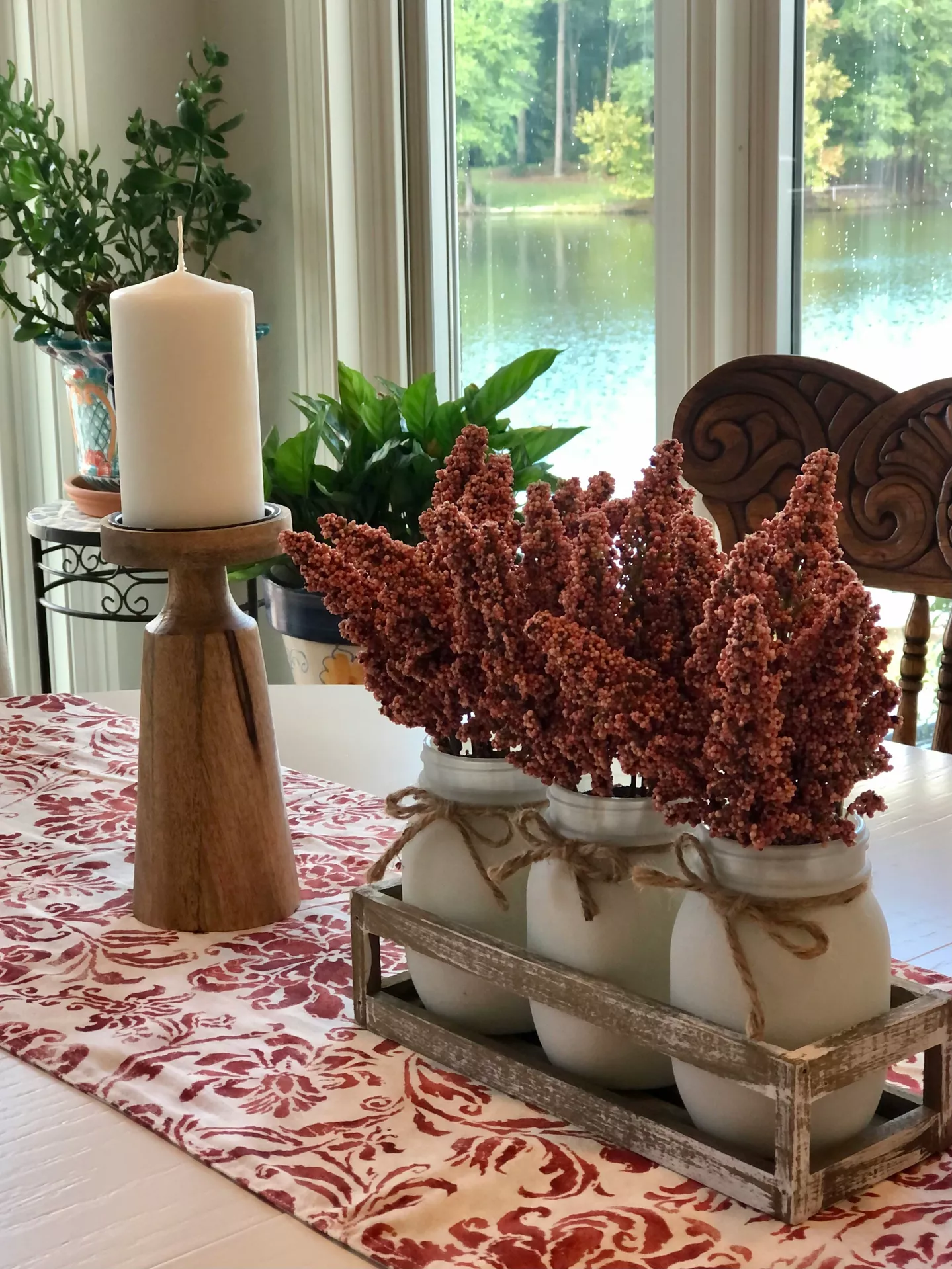 This image captures charming and cozy fall table decor ideas arranged neatly on a dining table with a red and white patterned damask runner. The main centerpiece consists of a rustic wooden crate holding three white frosted glass jars, each wrapped around the neck with a jute twine bow. The jars are filled with plush, reddish-brown stalks of dried sorghum that add a beautiful seasonal texture. To the left, a tall, thick wooden pillar candle holder stands proudly, supporting a single large unlit white candle. In the bright background, lush green potted houseplants sit near a large window that overlooks a scenic view of a calm lake and distant trees, completing the warm and natural autumnal look.