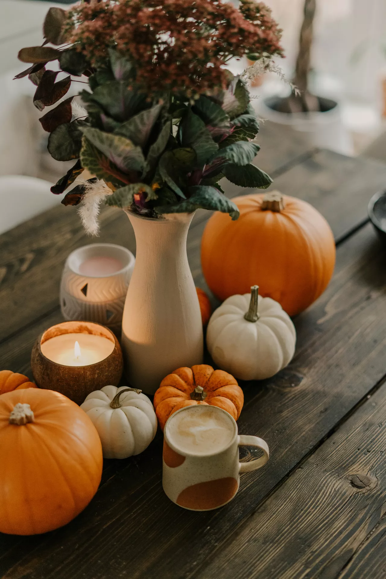 This image features inviting and cozy fall table decor ideas arranged on a dark, rustic wooden tabletop. The display includes an elegant collection of pumpkins, tea lights and flowers that bring a seasonal touch to the setting. In the center, a tall matte beige ceramic vase holds a dark floral bouquet with deep green and purple leaves alongside dusty pink blossoms. Clustered around the vase are various pumpkins in different sizes, including large classic orange pumpkins, small ribbed white pumpkins, and mini orange gourds. Adding to the warm ambiance are glowing candles, including a large tea light candle lit inside a natural coconut shell holder and a decorative white ceramic cut-out lantern. In the foreground, a ceramic mug filled with a warm latte with milk art sits ready, completing the comforting autumn atmosphere.
