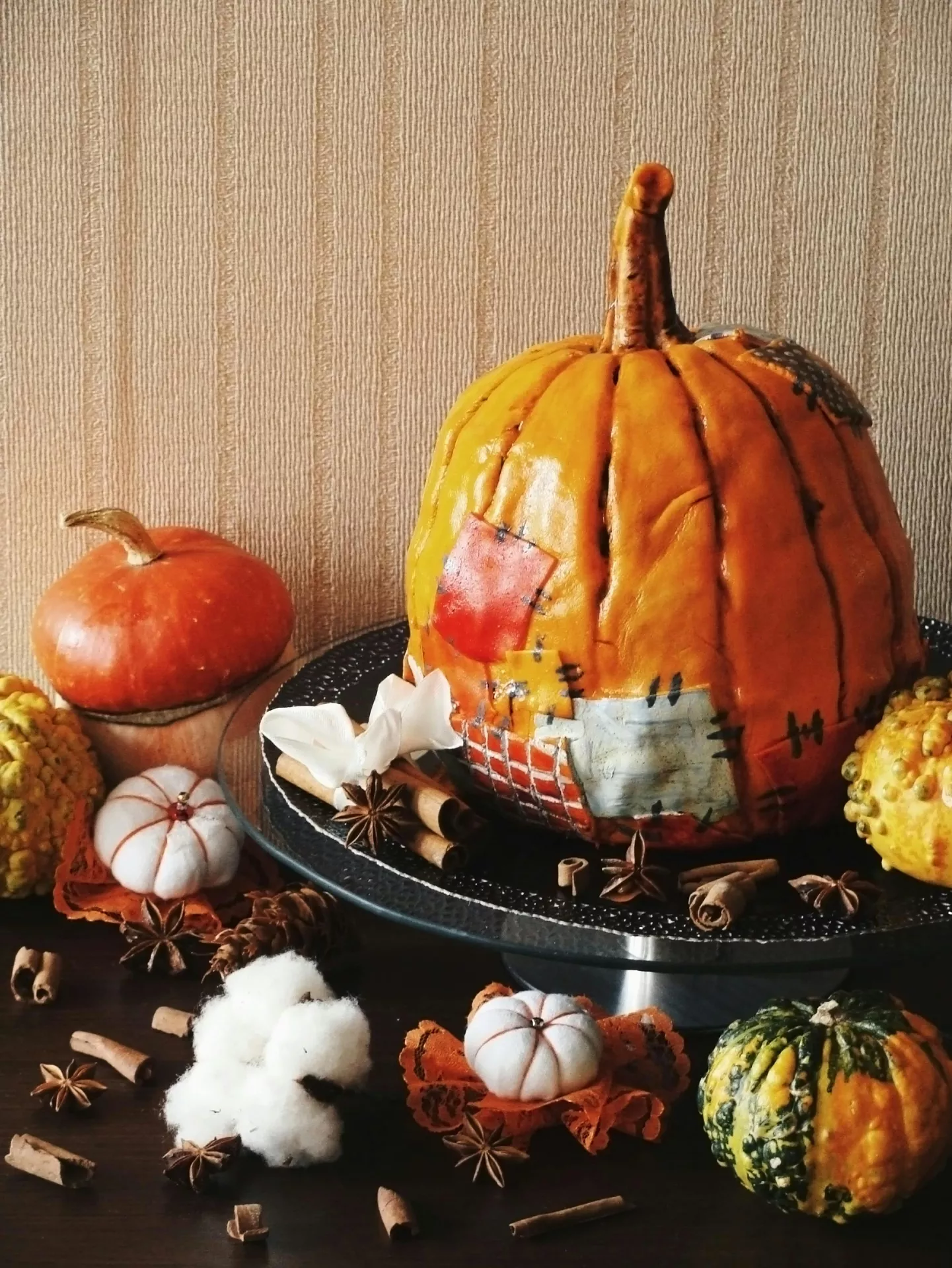 A large decorative orange pumpkin with patchwork designs rests on a black glass raised stand, serving as a center element for autumn styling. Around the large pumpkin, a small orange gourd, cinnamon sticks, star anise, and a white cotton-like flower are arranged on the tray. On the dark surface below the stand, additional festive elements are scattered, including a green and yellow striped gourd, tiny white fabric pumpkins, pinecones, more spices, and soft cotton pods against a neutral textured wall background. This arrangement offers creative fall table decor ideas featuring a pumpkin, cinnamon stick and cotton plant on lifted tray.