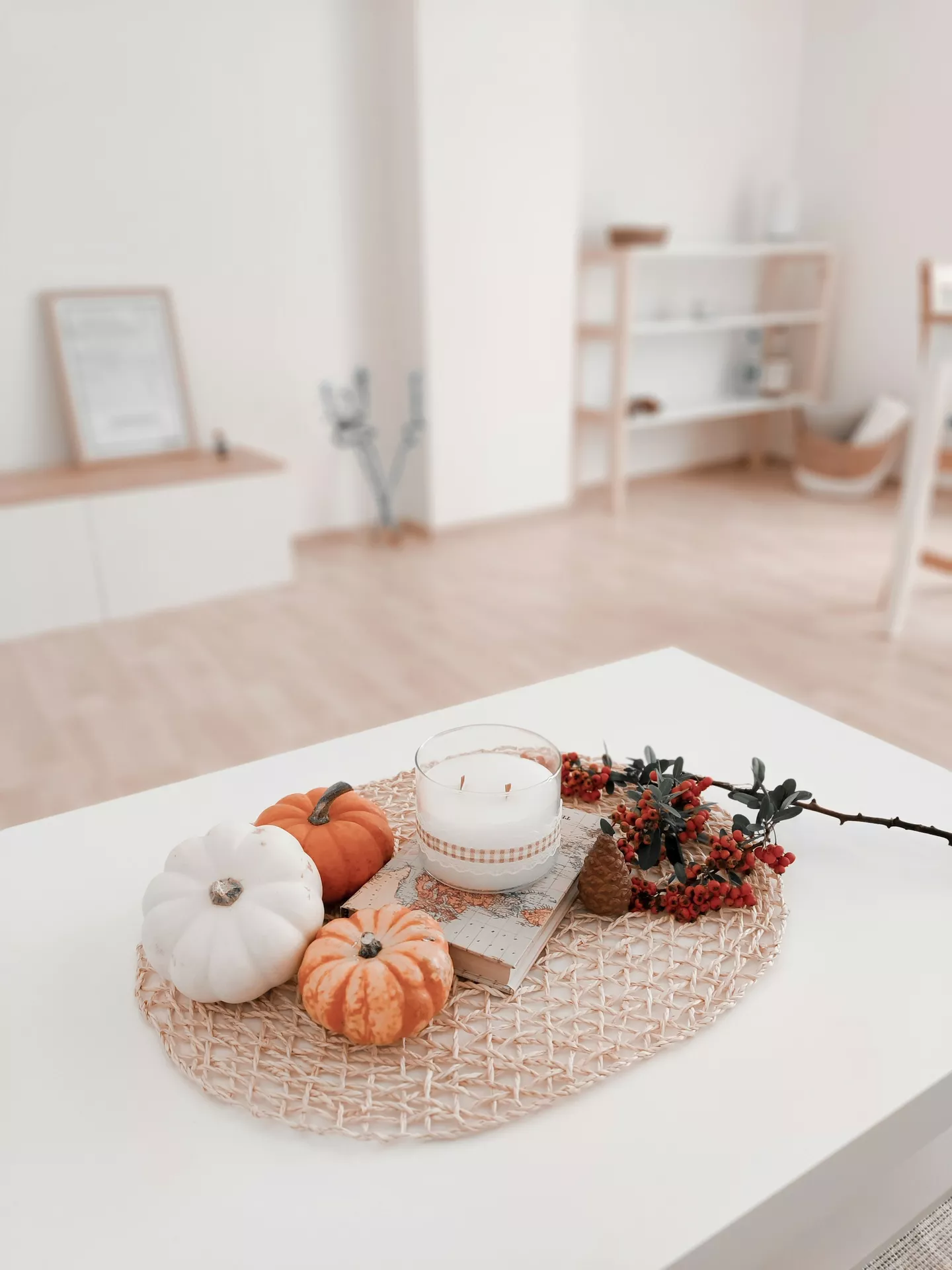 A bright and minimalist living room showcasing simple fall table decor ideas on a modern white table. A round woven placemat holds a cozy autumn arrangement featuring three small pumpkins, candle and red berry bush alongside a pinecone. The white glass candle sits on top of a small vintage book, perfectly complementing the clean and light interior space in the background.