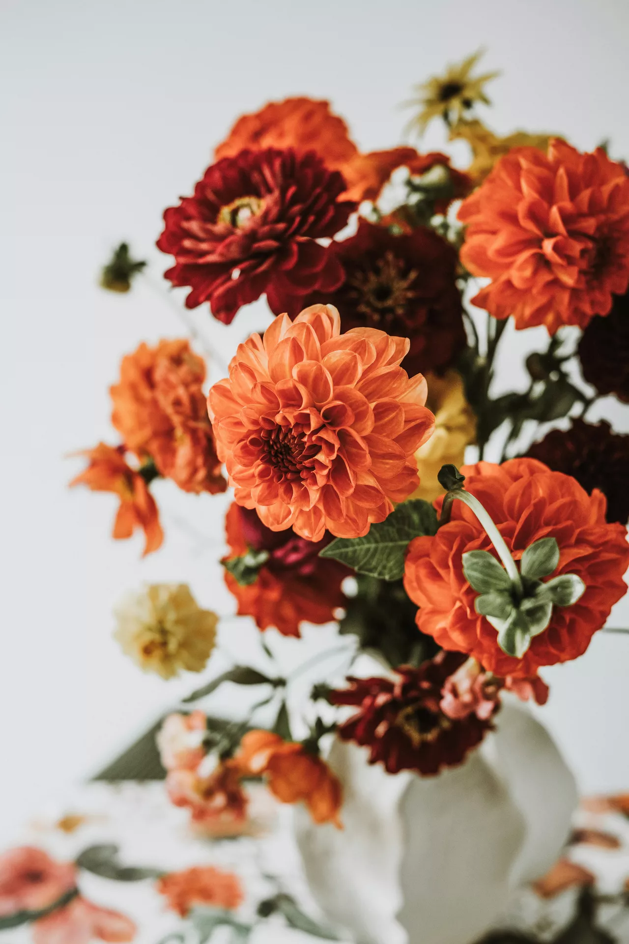 A beautiful close up of an autumn floral arrangement in a white vase featuring vibrant red and orange flowers such as dahlias against a bright background, providing elegant and natural fall table decor ideas for the season.
