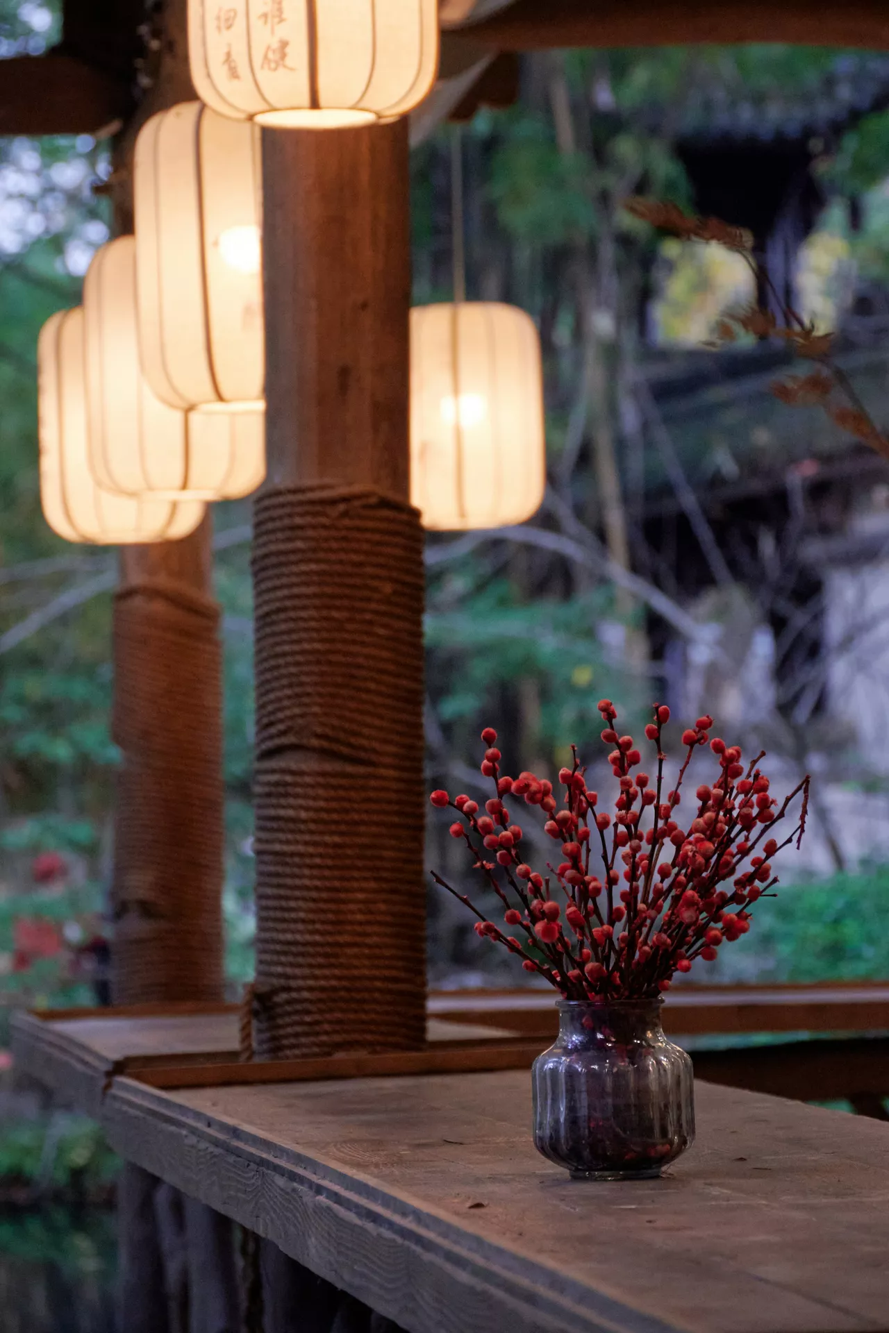 A rustic wooden outdoor table setting showcasing elegant fall table decor ideas. A small glass vase is filled with vibrant branches from red berry bushes, standing in front of thick rope-wrapped wooden pillars. Warm glowing paper lanterns hang above, creating a cozy and inviting autumn atmosphere against a lightly blurred natural background.