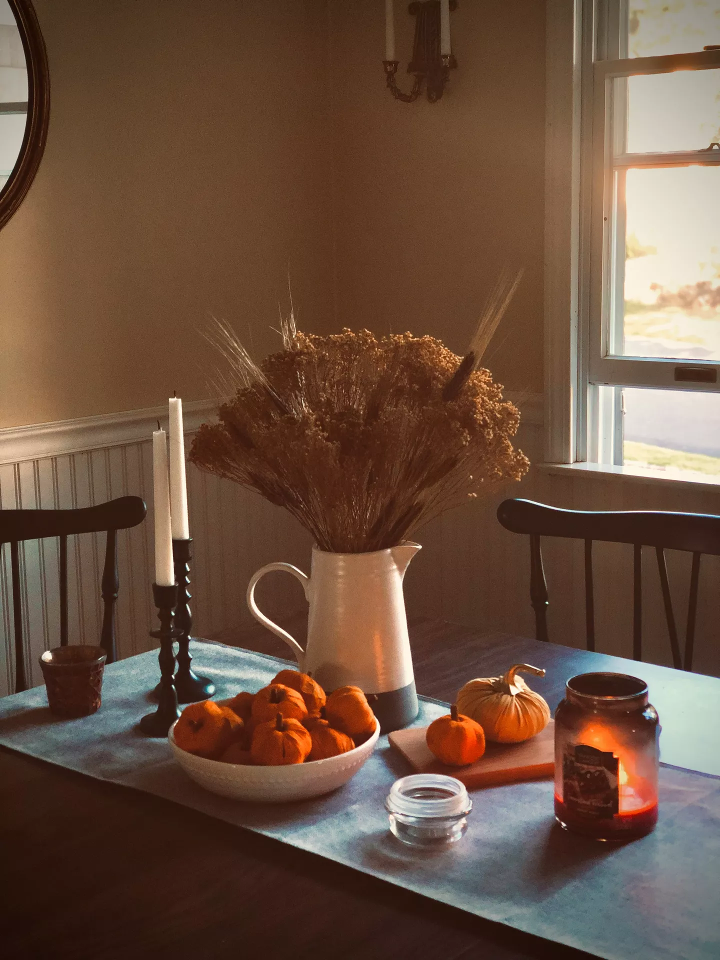 The file fall-table-decor-ideas-small-pumpkins-and-dried-flowers.jpg is a photograph of a dining table arrangement focused on autumn themes. The image displays a curated collection of fall table decor ideas on a wooden table with a textured blue table runner. The centerpiece is dominated by a white ceramic pitcher holding a tall, dense bouquet of small pumpkins and dried flowers, including wheat and various grasses. Adjacent to it, a white bowl is filled with numerous small pumpkins, and two other smaller individual small pumpkins and dried flowers sit on a small wooden board near a lit jar candle in a glass. The scene includes a pair of black candlestick holders with white tapers, and in the background, black wooden dining chairs are partially visible against beige walls with white wainscoting and a window with natural light on the right.