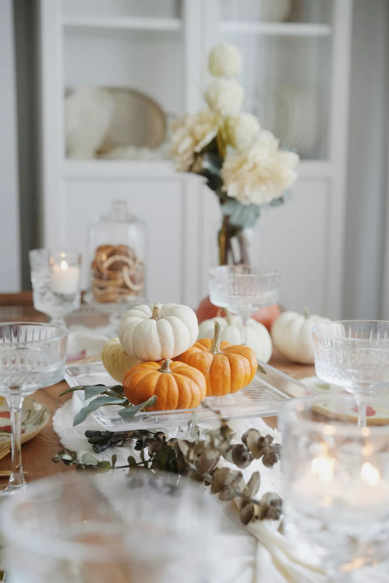 A bright and elegant dining room setting showcasing inspiring fall table decor ideas. The centerpiece features a mix of white and orange small pumpkins on a lifted tray made of clear acrylic, resting on a softly textured white fabric runner. The festive arrangement is surrounded by dried eucalyptus branches, vintage crystal glassware, and softly glowing candles. In the lightly blurred background, a white display cabinet, a glass cloche filled with dried citrus slices, and a vase with delicate white autumn flowers complete the sophisticated seasonal aesthetic.