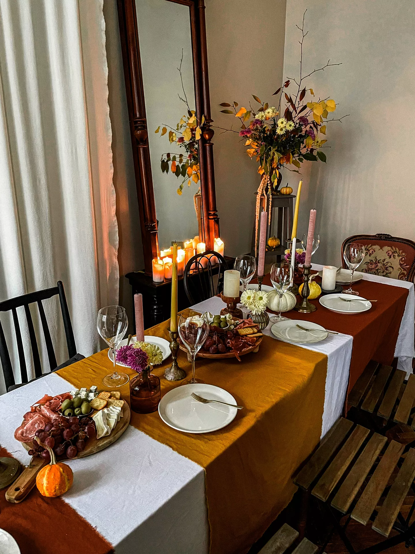 A richly decorated dining table set for an autumn celebration, showcasing a multitude of fall table decor ideas. The table features layered runners in white, gold, and burnt orange and is laden with place settings with white dishes, gold cutlery, and various wine glasses. The central decorations consist of numerous small vases and candles. Small vases filled with autumnal flowers like chrysanthemums and branches are interspersed with diverse taper (gold, pink, ivory) and pillar candles in metallic and wooden holders. Two wood charcuterie boards are piled with snacks, and small pumpkins are scattered along the table. In the background, a large wooden mirror reflects the warm candlelight, with a tall floral arrangement of autumn leaves and branches and chairs on either side. The lighting is warm and ambient.