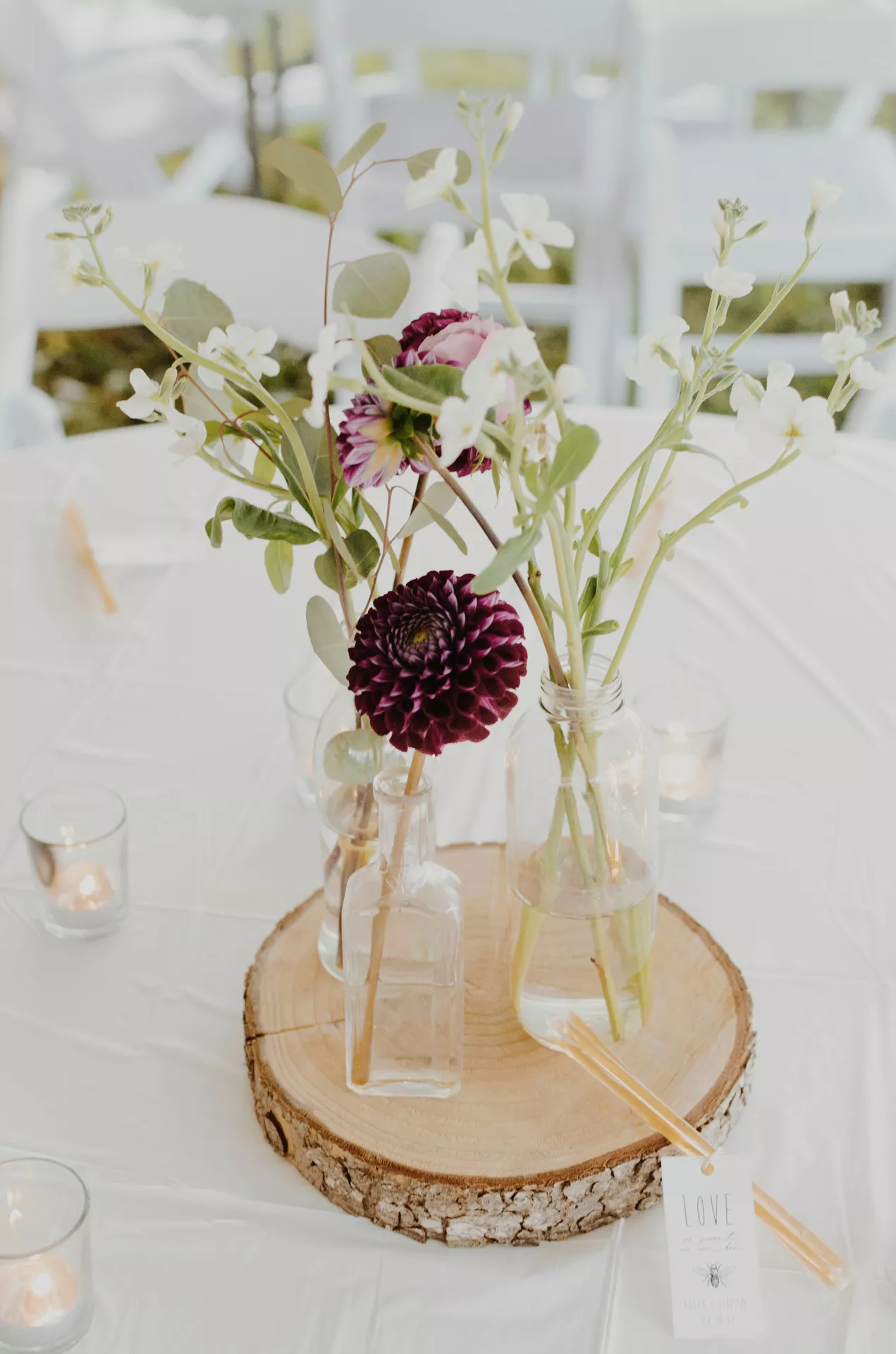 A bright and rustic event centerpiece providing elegant fall table decor ideas. The arrangement features clear glass small vases on tree trunk slice, holding a delicate mix of deep purple dahlias, small white blooms, and green foliage. The wooden base rests on a crisp white tablecloth and is surrounded by small, warmly glowing votive candles in clear glass holders. Resting gently on the bark edge of the wood slice are honey sticks and a small white tag displaying the word LOVE and a tiny bee illustration, set against a softly blurred background of white folding chairs.