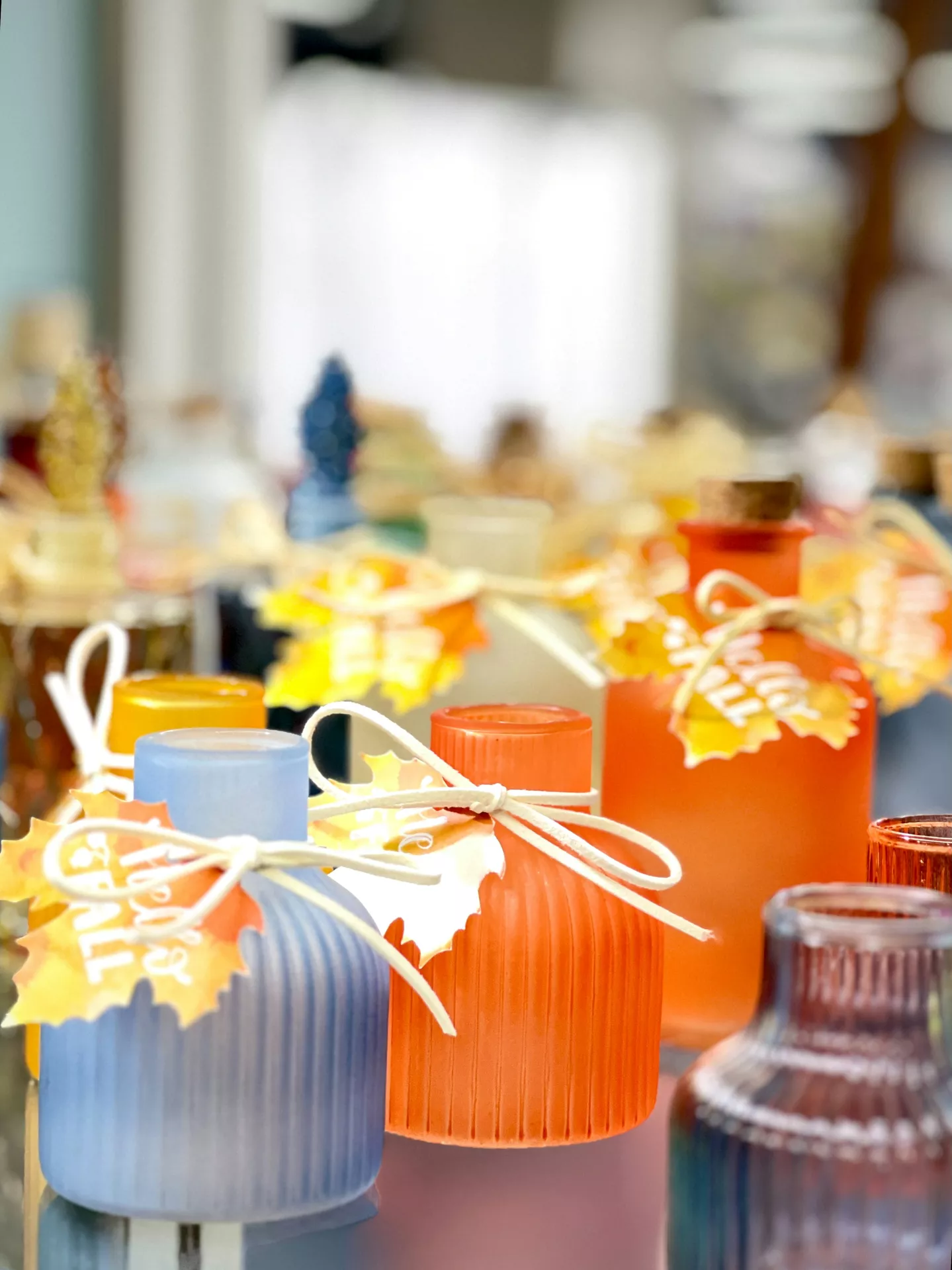 A close up view showcasing colorful fall table decor ideas. In the foreground, a ribbed light blue glass vessel and a bright orange tea light holder with leaf decoration are tied around the neck with a white string bow. The orange and yellow leaf shaped paper tags have the words Hello Fall written on them. The background features a softly blurred arrangement of similar festive frosted glass bottles in various warm autumn colors, creating a cozy seasonal aesthetic.