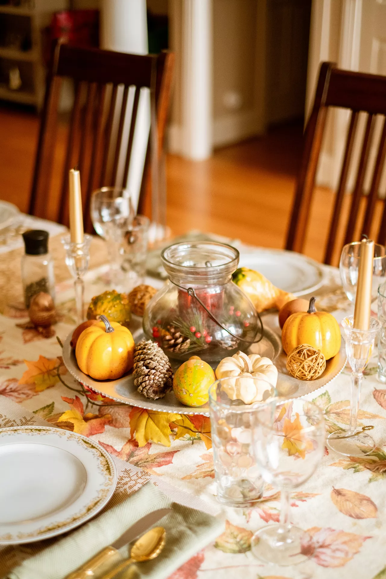 A warm dining table setting showcasing beautiful fall table decor ideas. The centerpiece features a round silver tray with pumpkins, candle and pine cones, surrounded by small gourds and woven decorative spheres. The table is elegantly arranged with a fall leaf patterned tablecloth, gold-rimmed white plates, gold flatware, clear crystal glassware, and tall gold taper candles, set against a background of wooden dining chairs and a hardwood floor.