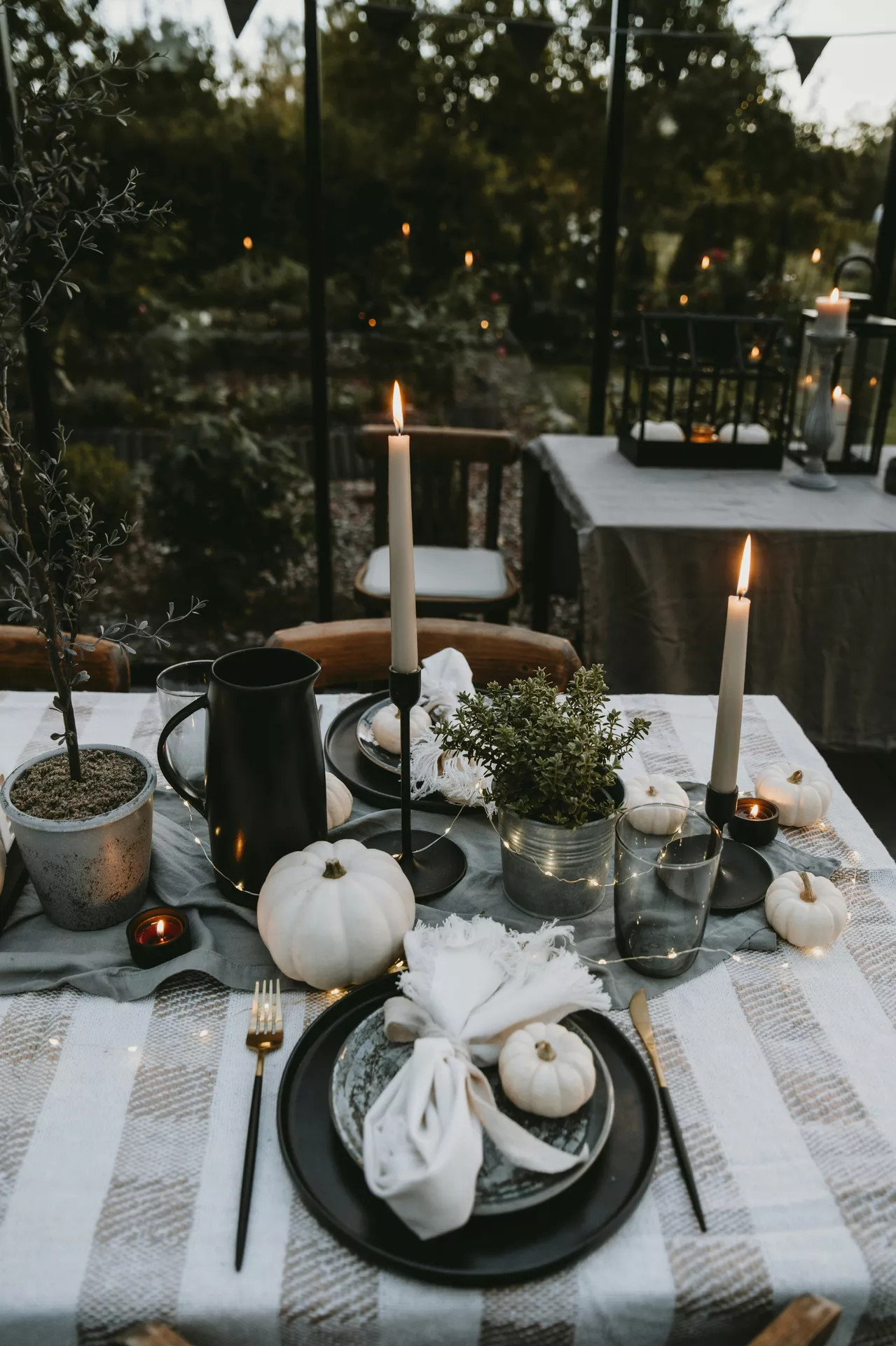 An elegant outdoor evening dining setting showcasing beautiful fall table decor ideas. The table is laid with a lightly striped tablecloth and a soft grey fabric runner, decorated with a modern black pitcher, small potted greenery, and a glowing arrangement of white pumpkins, candles and fairy lights. The rustic place settings feature dark plates topped with loosely tied white napkins and miniature pumpkins, complemented by black and gold flatware against a moody, softly lit garden background.