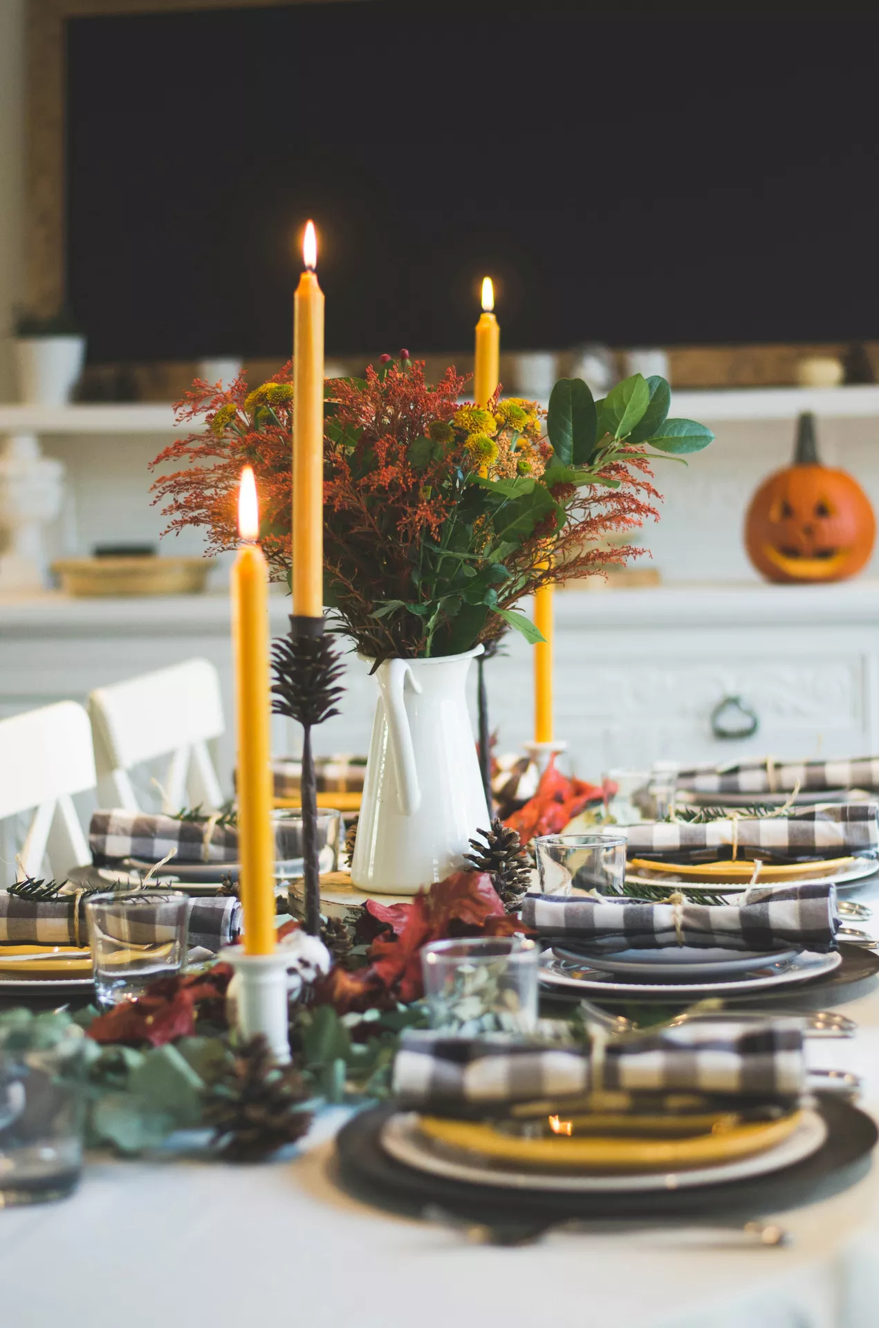 A beautifully set dining table showcasing inspiring fall table decor ideas. A white pitcher filled with seasonal flowers and foliage serves as the centerpiece, surrounded by tall glowing yellow candles, autumn green and pine cones scattered along the table runner. The place settings feature black chargers, yellow plates, and black and white plaid napkins tied with twine. In the softly blurred background, white dining chairs and a carved jack-o-lantern on a white sideboard complete the festive holiday atmosphere.