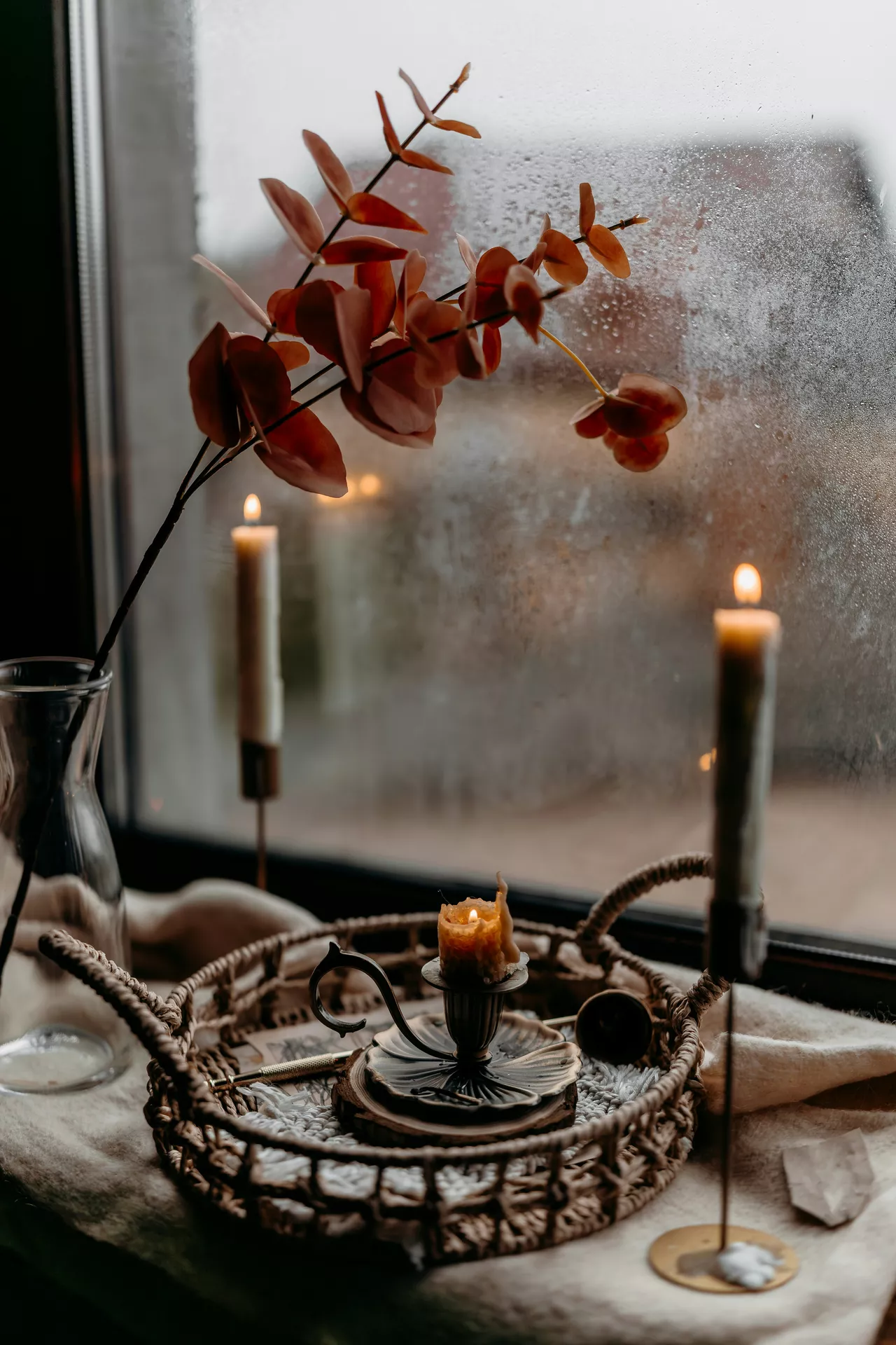 Fall tray decor ideas featuring candles on candle holders and dry flowers arranged on a woven tray by a misty window, creating a warm and cozy autumn display with soft candlelight and natural seasonal accents.
