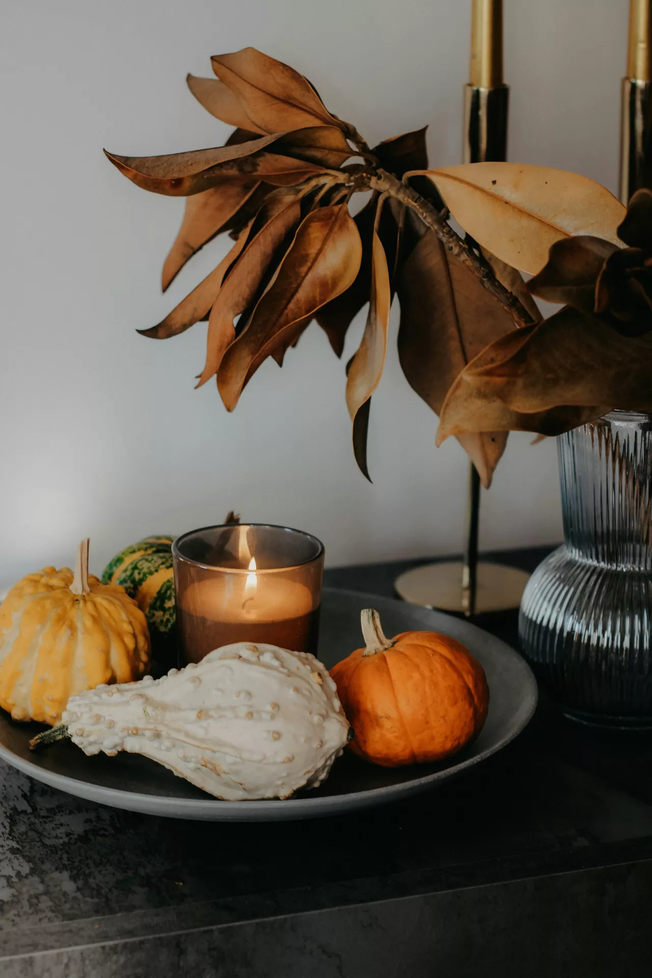 Fall tray decor ideas featuring colorfull different pumpkins arranged on a decorative tray with a glowing candle, dried magnolia leaves in a glass vase, and elegant candle holders for a warm and cozy autumn centerpiece.