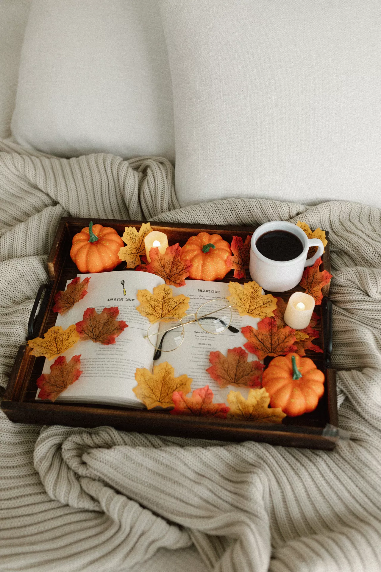 Fall tray decor ideas featuring Leaves & Mini Pumpkins arranged on a rustic wooden tray with colorful autumn leaves, an open book, reading glasses, glowing candles, and a warm cup of coffee, creating a cozy fall reading nook on a soft knitted blanket.