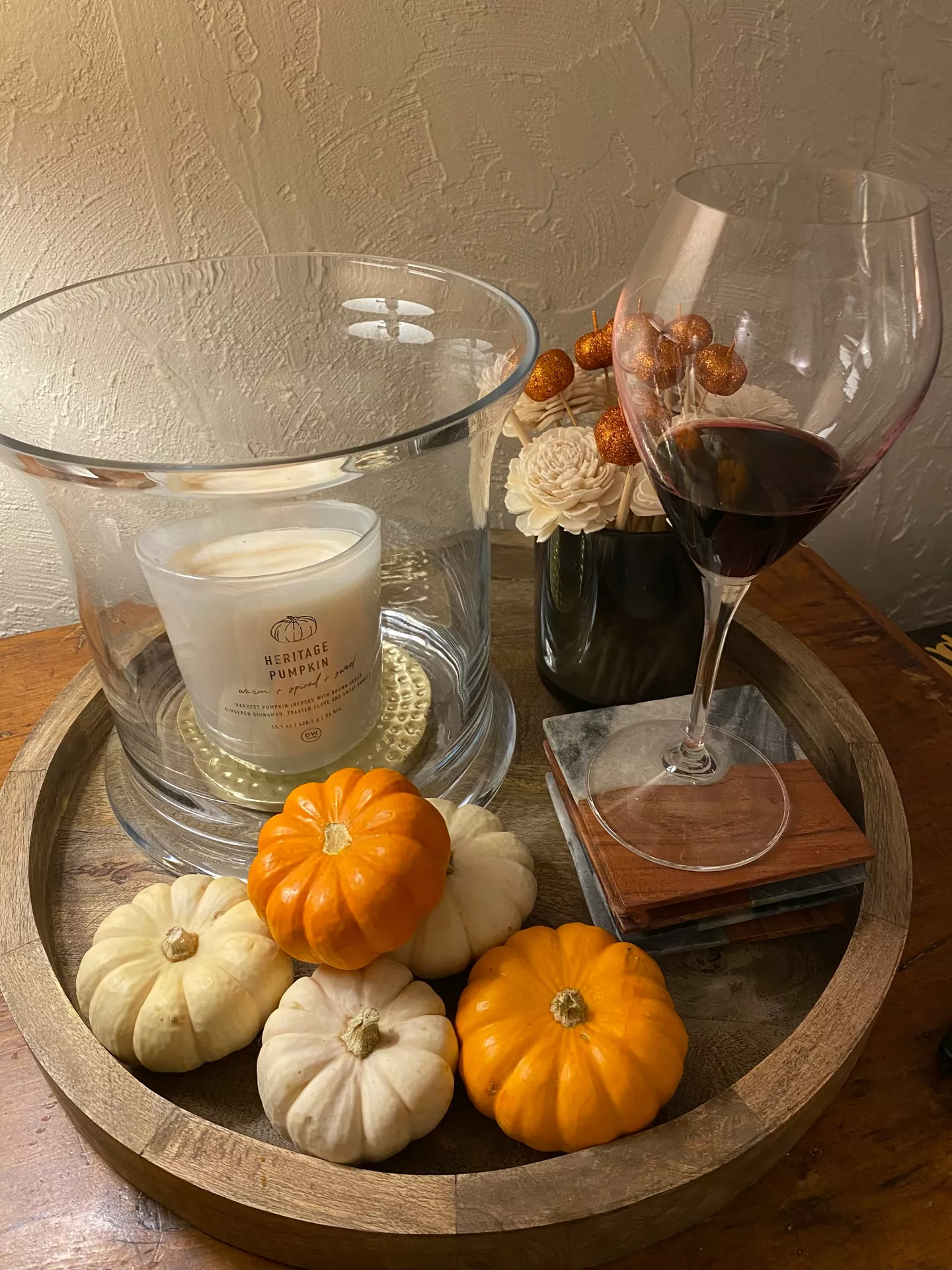 Fall tray decor ideas featuring Mini Pumpkins, Candle & Dried Flowers arranged on a round wooden tray with orange and white mini pumpkins, a large glass hurricane candle, dried floral accents, a glass of red wine, and rustic coasters for a warm and inviting autumn display.