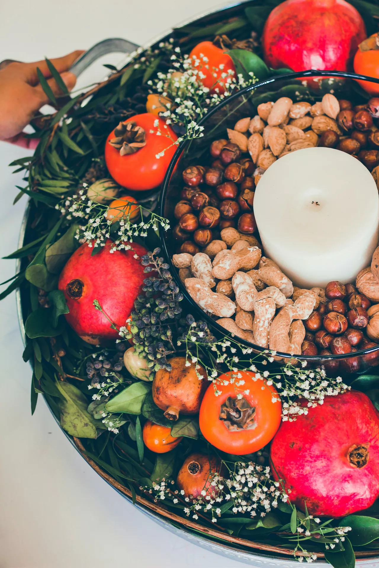 Fall tray decor ideas featuring Pomegranates, Persimmons & Nuts arranged in a large decorative tray with lush greenery, vibrant pomegranates, ripe persimmons, assorted nuts, delicate white flowers, and a pillar candle for a rich and abundant autumn centerpiece.