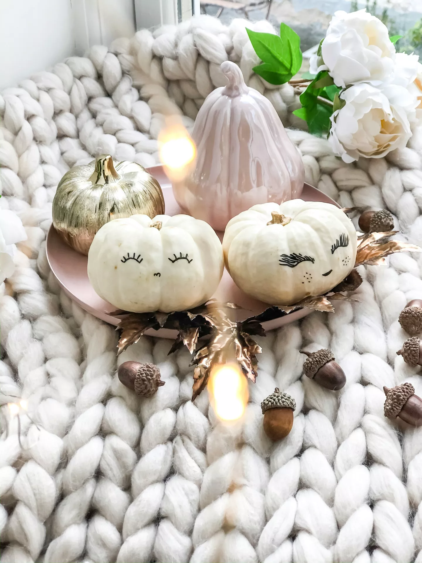Fall tray decor ideas featuring white pumpkins with faces arranged on a blush pink tray alongside a gold pumpkin, a ceramic pear, metallic leaves, scattered acorns, cozy knit blankets, and soft fairy lights for a charming and playful autumn display.