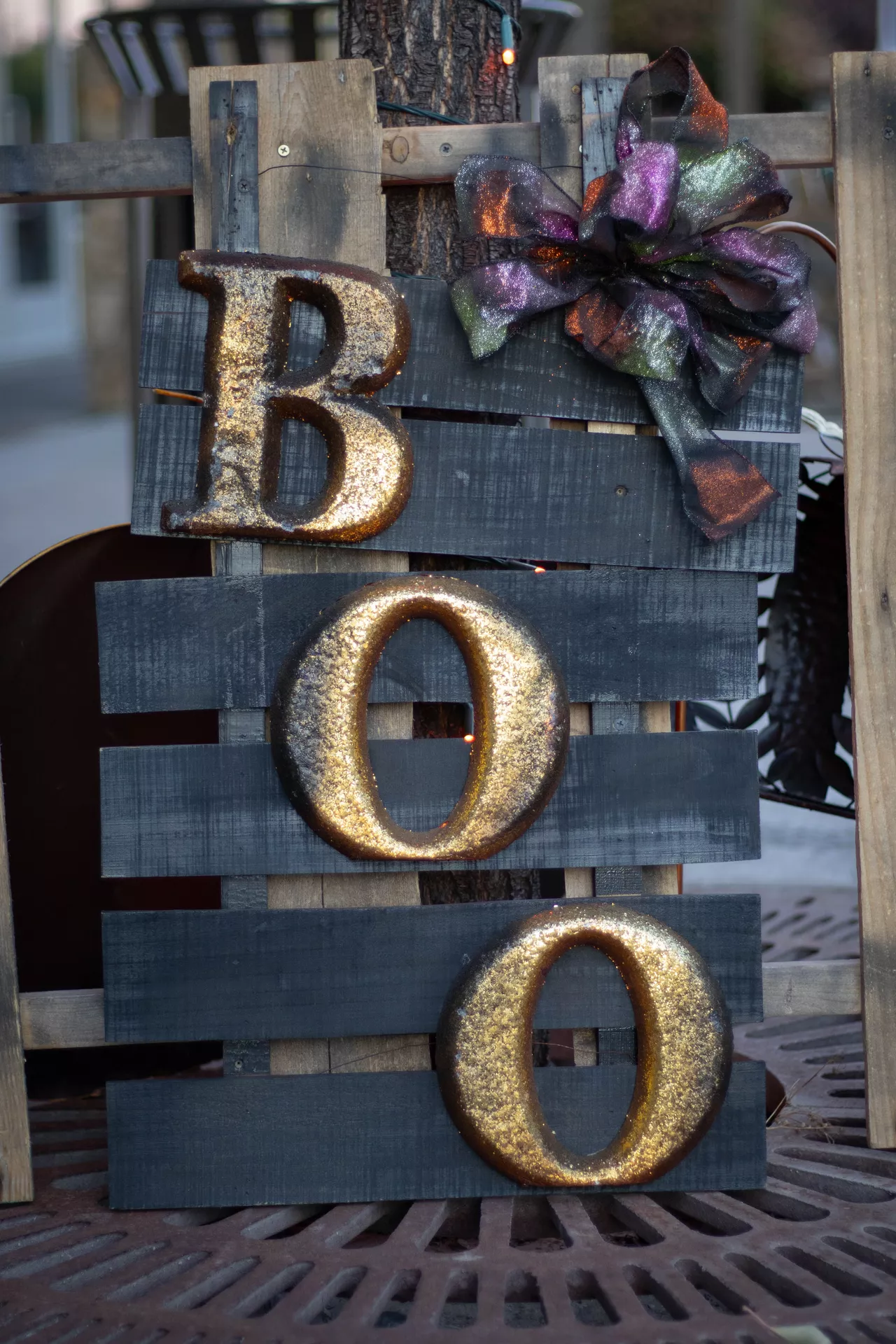 Rustic Boo Sign made from a painted wooden pallet with glittery gold letters, finished with a shimmering black and purple bow for a festive display. This charming outdoor decoration is perfect for Halloween Decor Ideas, adding a spooky yet welcoming touch to a front porch, garden, or entryway.