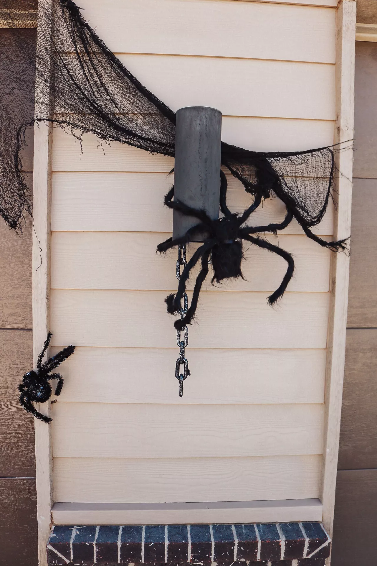 Large black decor spiders attached to a front porch column with spooky spider web fabric and hanging chain, creating a creepy outdoor display full of Halloween Decor Ideas. These oversized decor spiders add a dramatic haunted house look to an entryway or front porch.