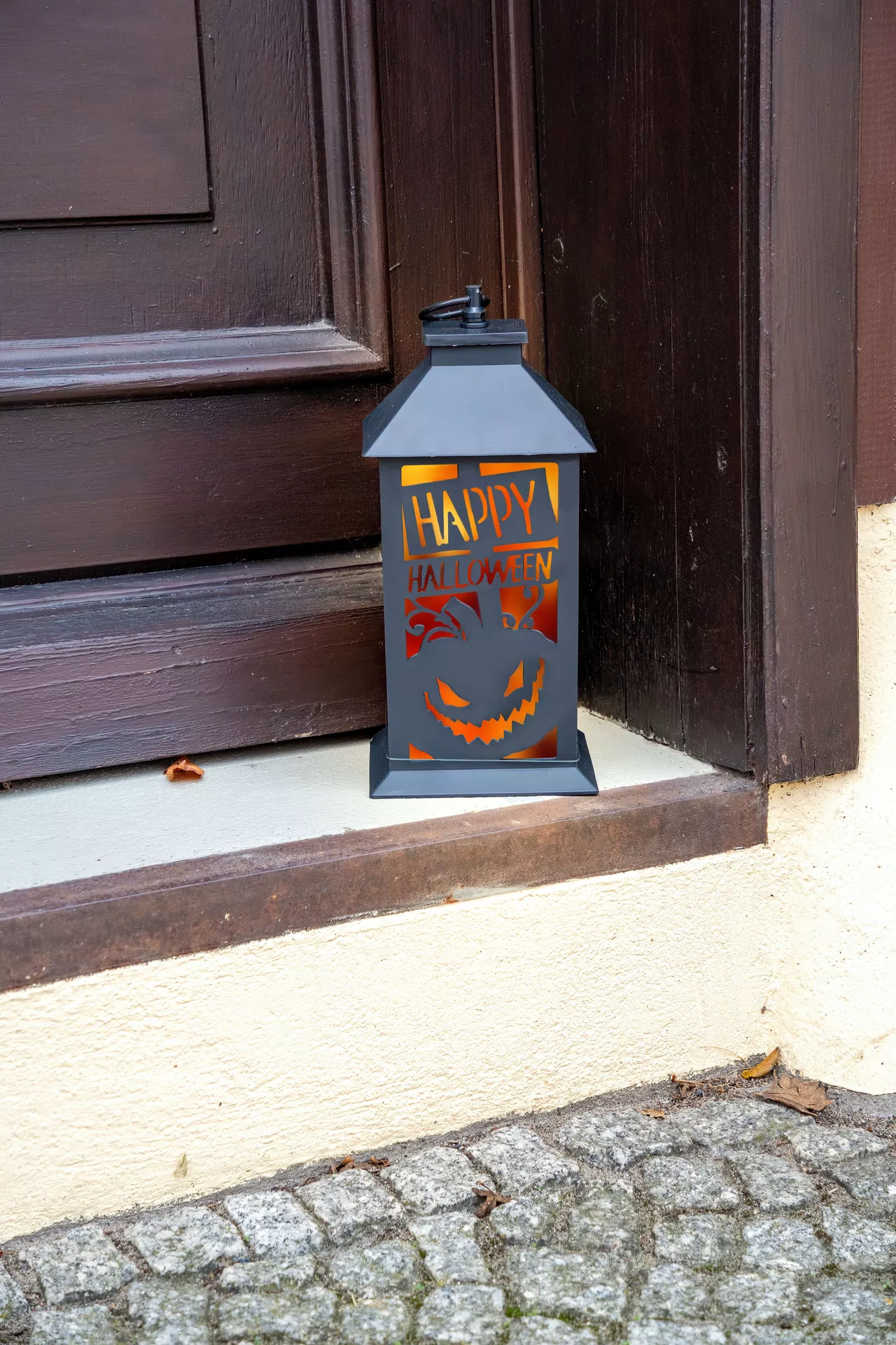 Black freestanding lantern with a glowing orange interior, featuring a carved Happy Halloween message and jack-o'-lantern design, displayed beside a front door. This festive accent is a great addition to Halloween Decor Ideas, adding a warm and spooky touch to a porch, doorstep, or entryway.