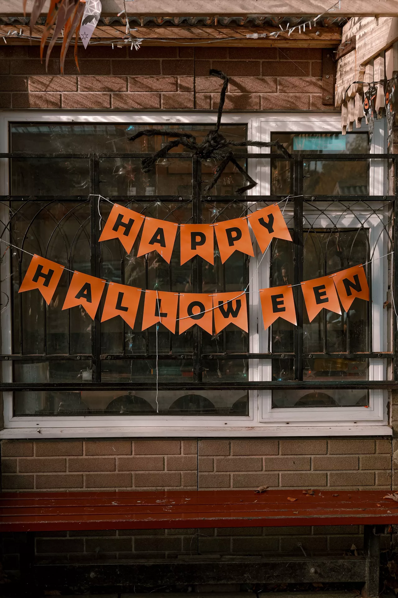 Orange happy halloween garland hanging across a porch window with a large black spider decoration above it, creating a fun and spooky outdoor display. This festive setup offers simple Halloween Decor Ideas, and the happy halloween garland adds a bold seasonal touch to a front porch or patio.