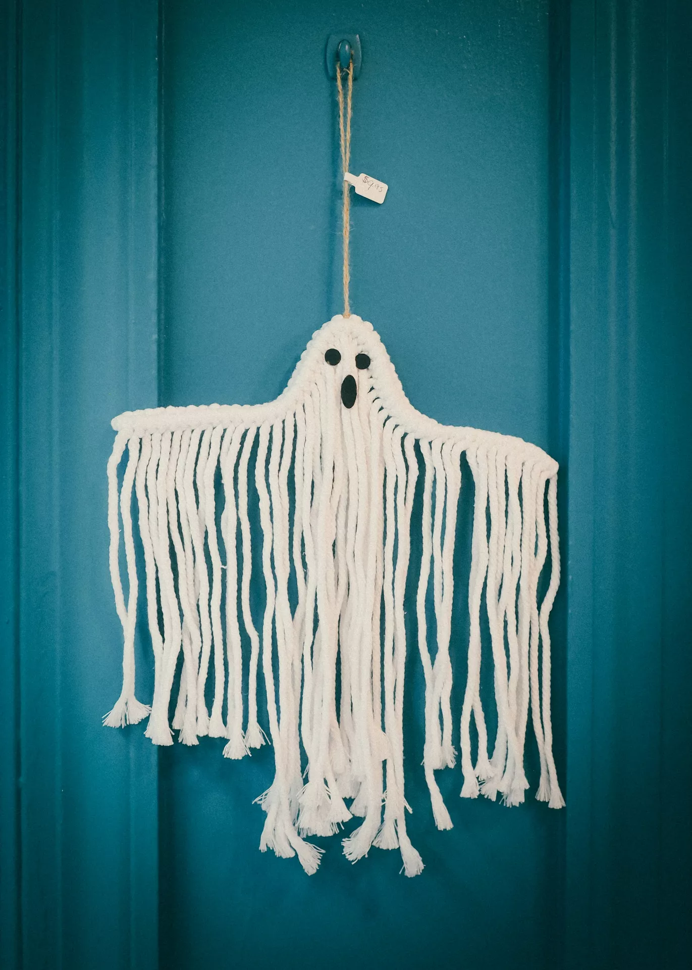 White makramé ghost wall hanging with long fringe and black felt eyes displayed on a teal front door, creating a soft handmade look for festive Halloween Decor Ideas. This charming makramé ghost adds a cozy boho touch to seasonal porch or entryway decorations.