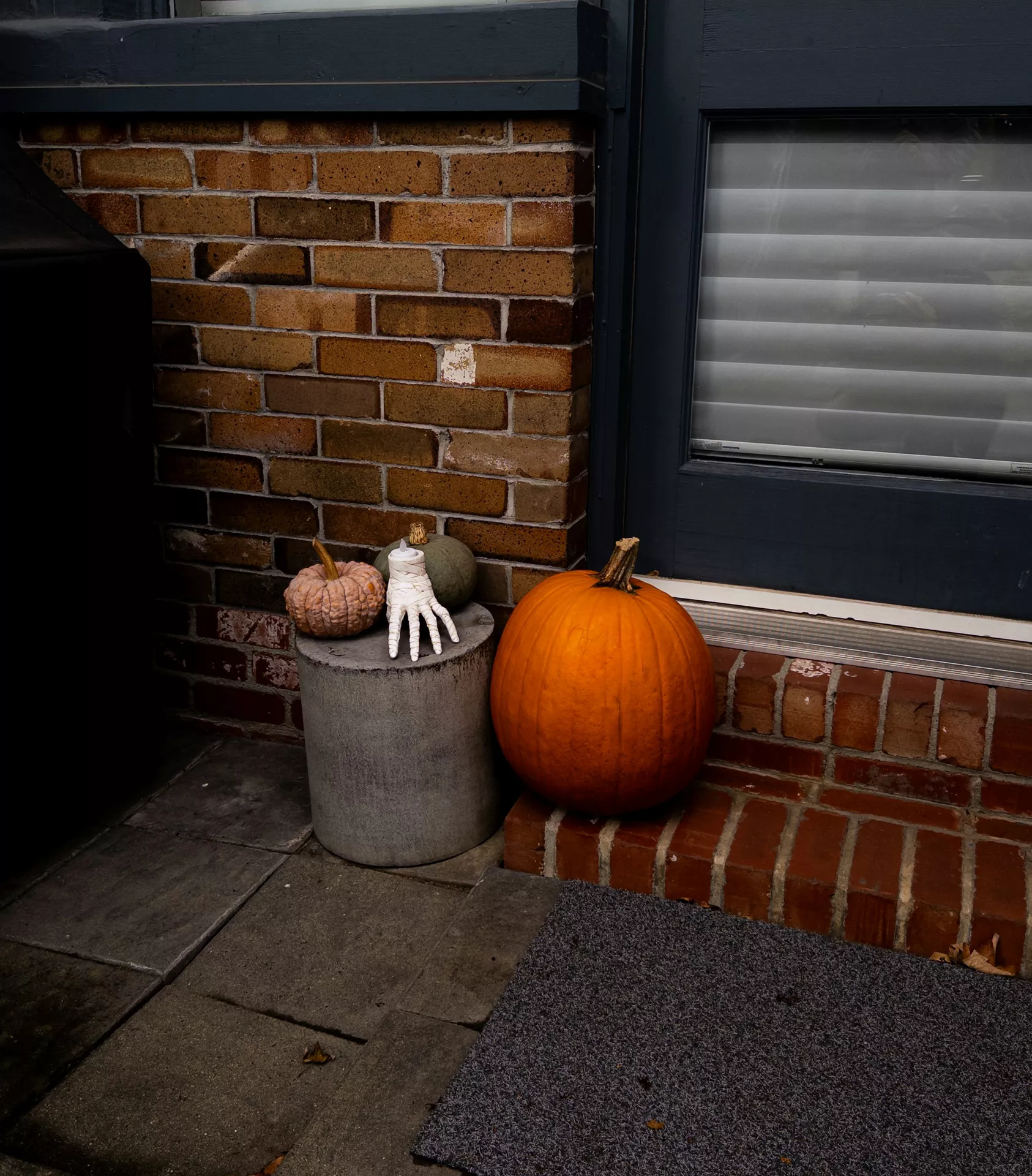 Outdoor Halloween display featuring a large orange pumpkin beside smaller decorative gourds and Thing from the show Wednesday resting on a concrete pedestal. Set against a brick wall and dark window frame, this simple arrangement offers creative Halloween Decor Ideas with pumpkin and thing from the show wednesday for a modern front porch.