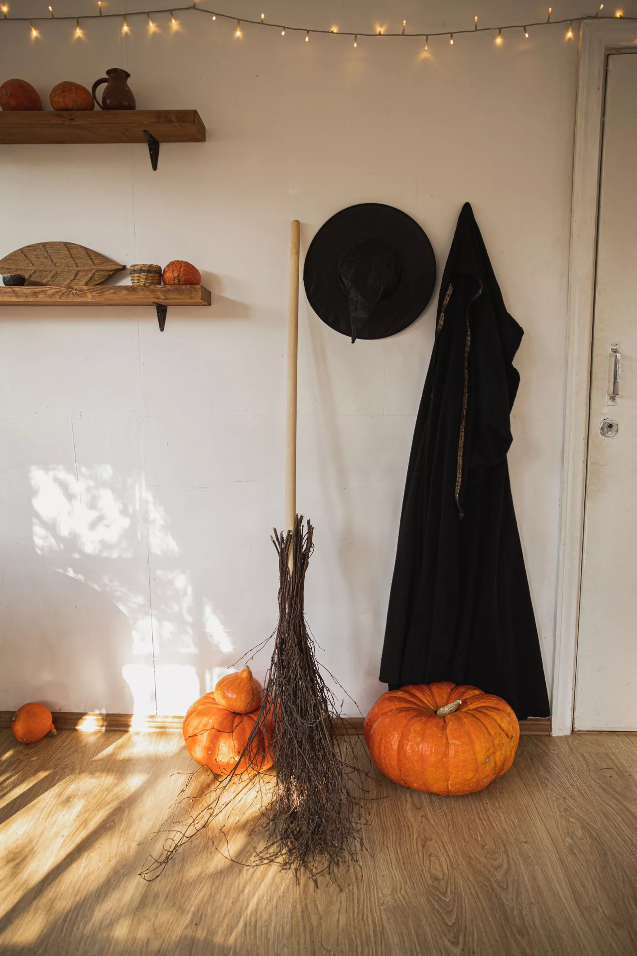Cozy Halloween setup featuring a rustic witch broom, black witch hat, hanging cloak, and large pumpkins arranged against a bright wall with wooden shelves and string lights. This simple display blends seasonal charm with classic witch decor, making it a stylish source of Halloween Decor Ideas for an inviting indoor space.