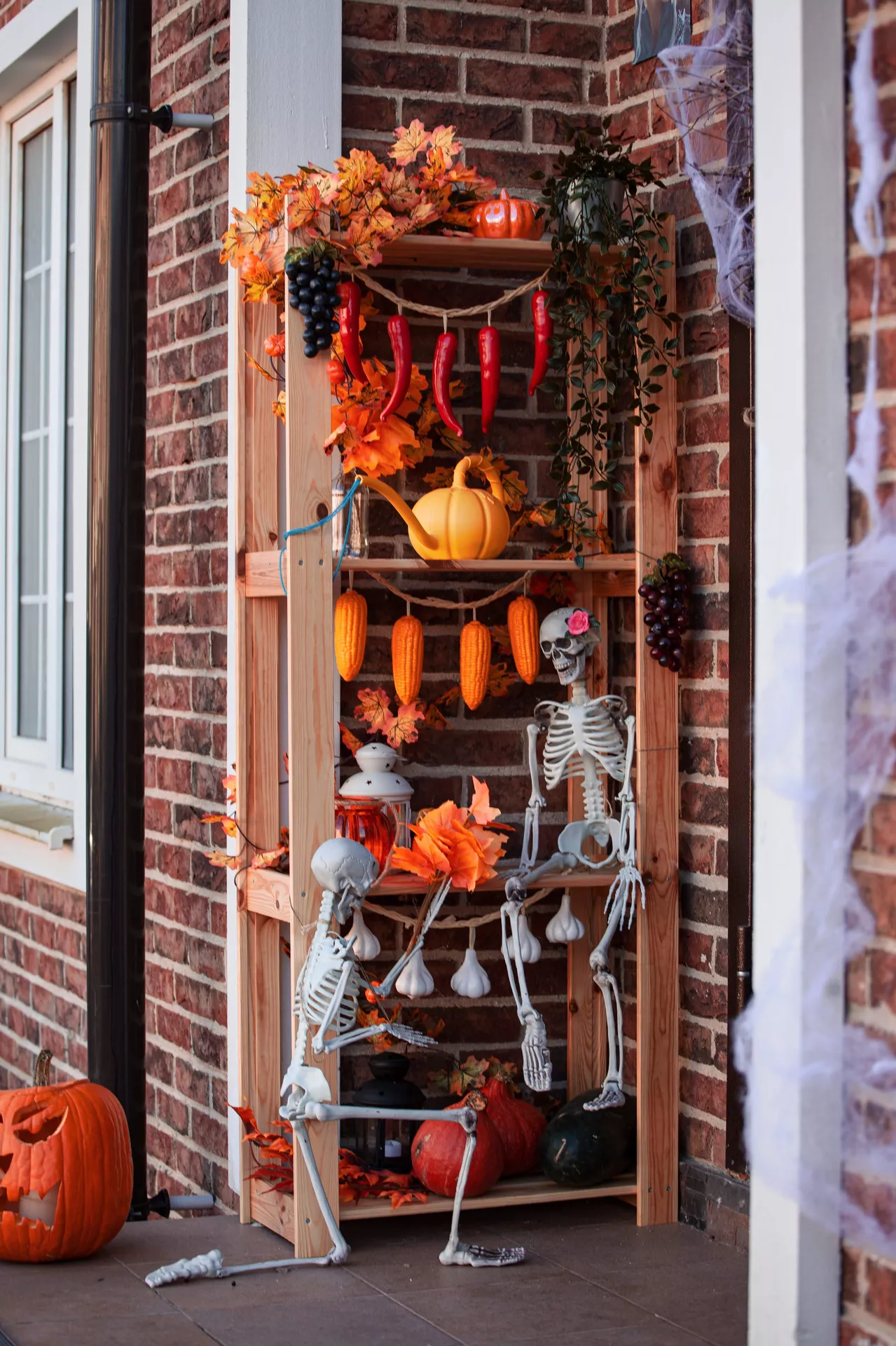 Halloween Decor Ideas featuring a wooden shelf with Halloween elements, including skeleton figures, pumpkins, autumn leaves, hanging garlic and corn, grape clusters, lanterns, and festive decorations arranged on a cozy front porch with seasonal fall accents.