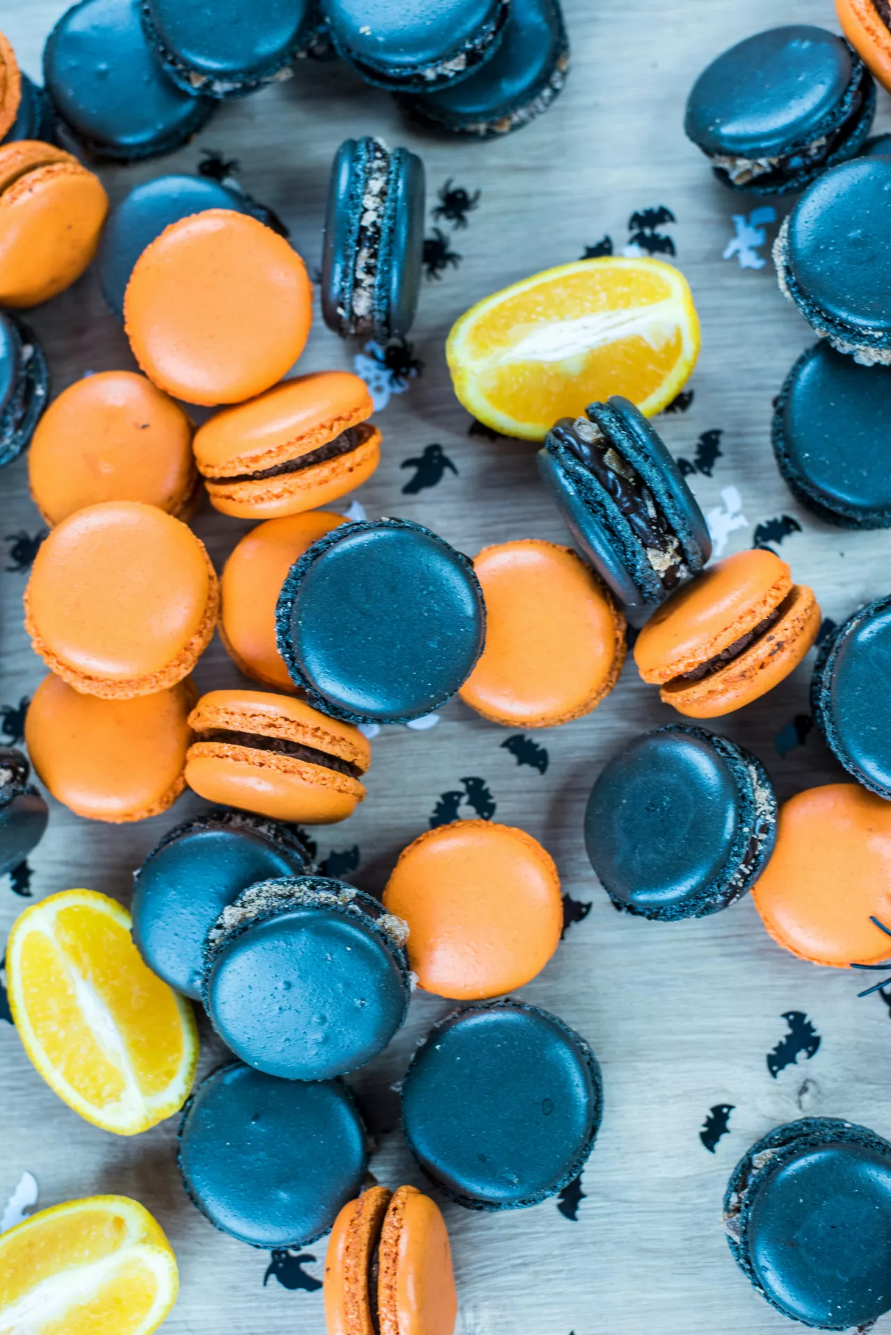 Halloween Party Food Ideas featuring black and orange macarons arranged with fresh orange slices and spooky Halloween confetti, creating a festive dessert display perfect for a stylish Halloween celebration.
