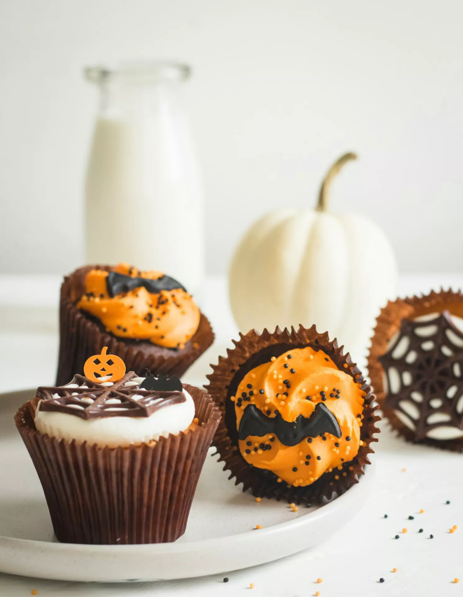 Halloween Party Food Ideas featuring cupcakes with pumpkin and bat decor, including chocolate cupcakes topped with orange frosting, spiderweb icing, Halloween sprinkles, and spooky decorations, styled beside a white pumpkin for a festive dessert display.